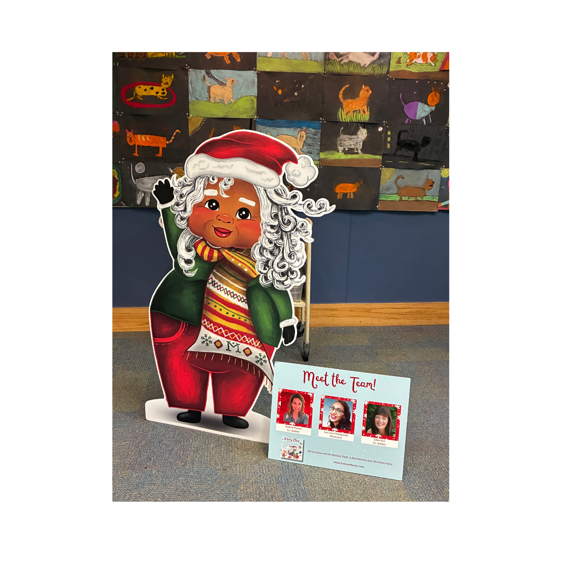 a five foot tall cut out of Mrs. Claus, Merry, stands next to a Meet the Team poster featuring the co-authors and the illustrator.