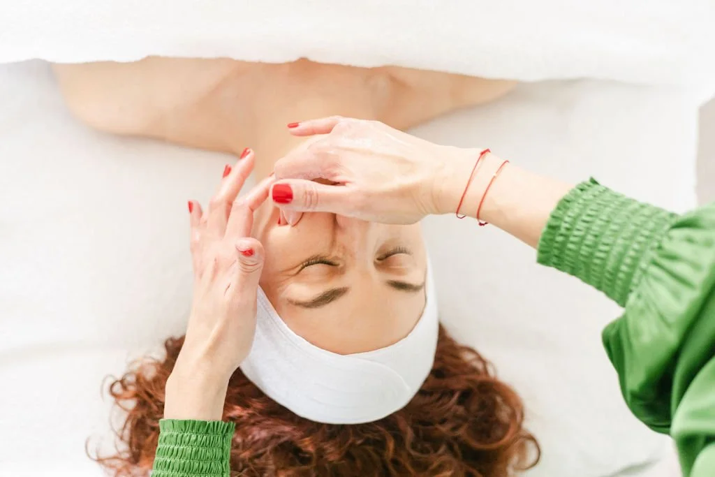 Woman receiving a facial massage at spa, lying down with eyes closed, wearing a white headband, with a massage therapist in green sleeve applying pressure to her forehead.
