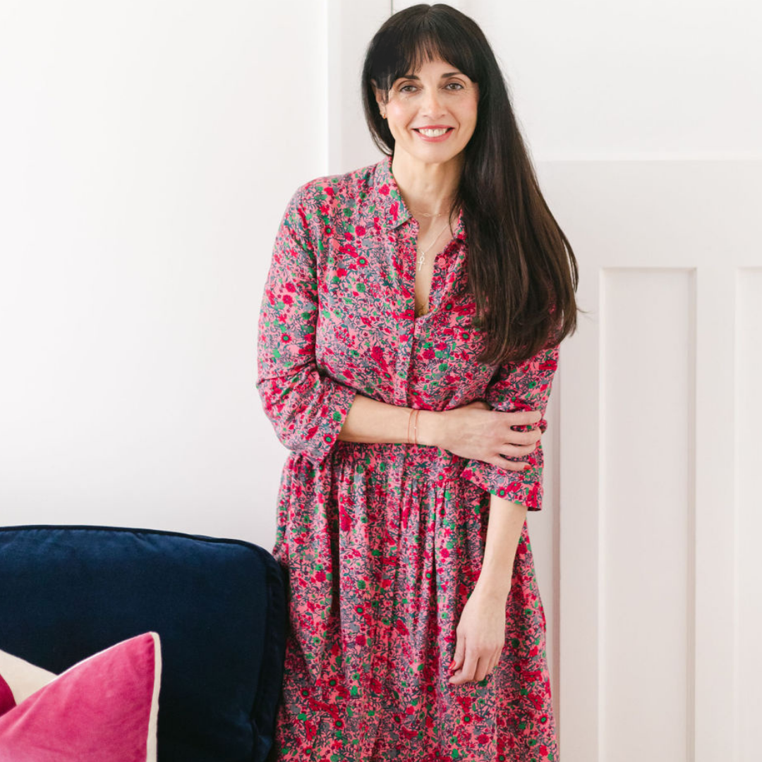 A woman with long dark hair and a bright smile, wearing a pink floral dress, standing near a white wall with a navy blue chair and pink pillows in the foreground.