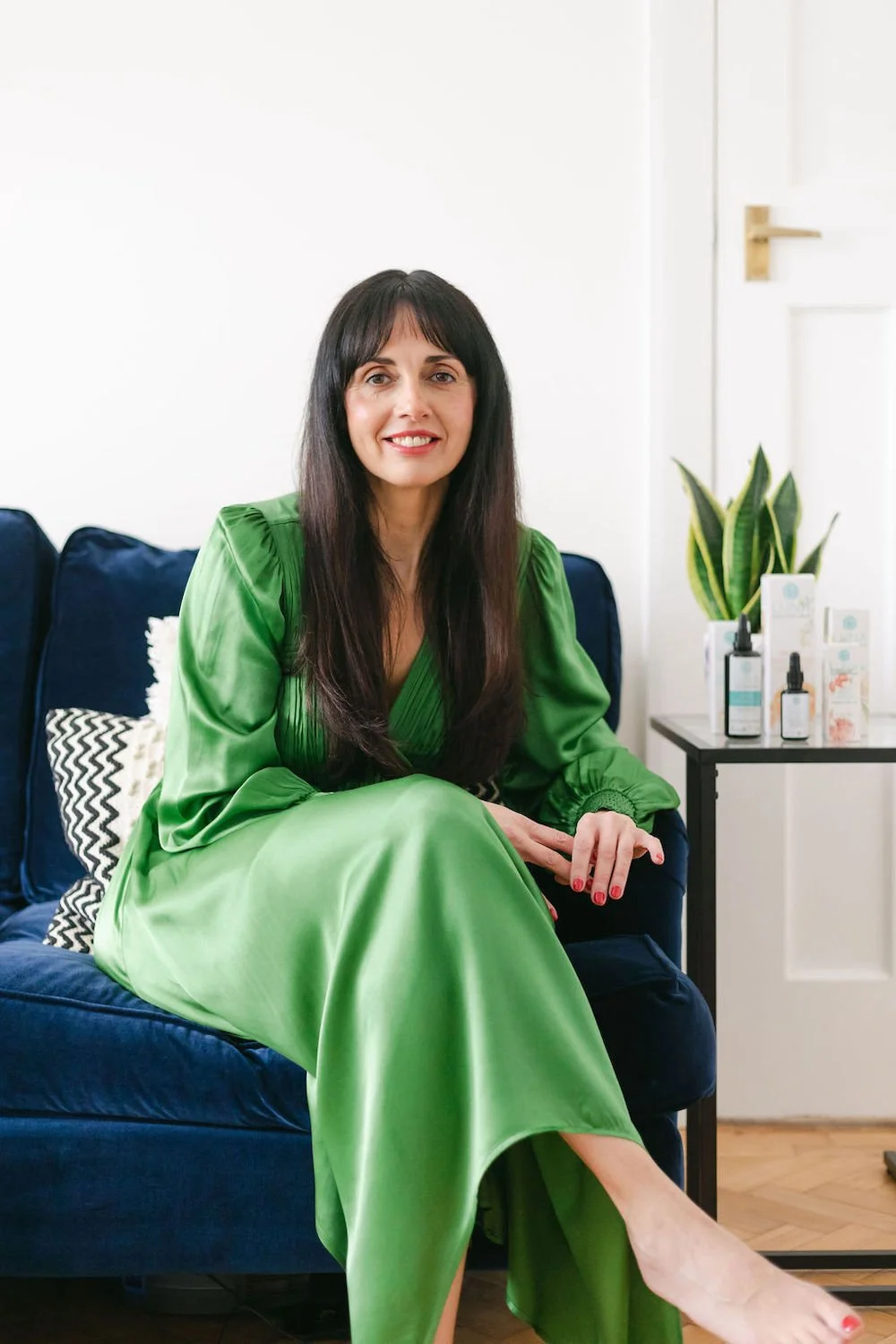 Woman with long dark hair sitting on a navy blue sofa, wearing a bright green satin dress, in a room with white walls and a small table with skincare products and a plant in the background.