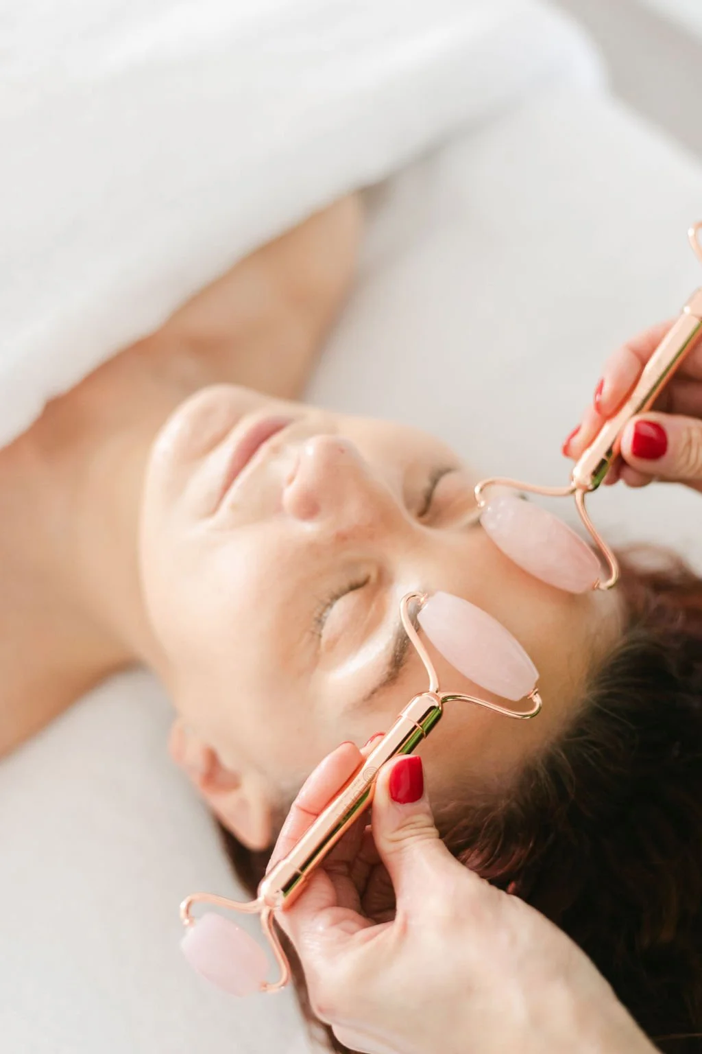 A woman with closed eyes receiving a facial treatment using a rose quartz roller on her face, lying down with a relaxed expression.