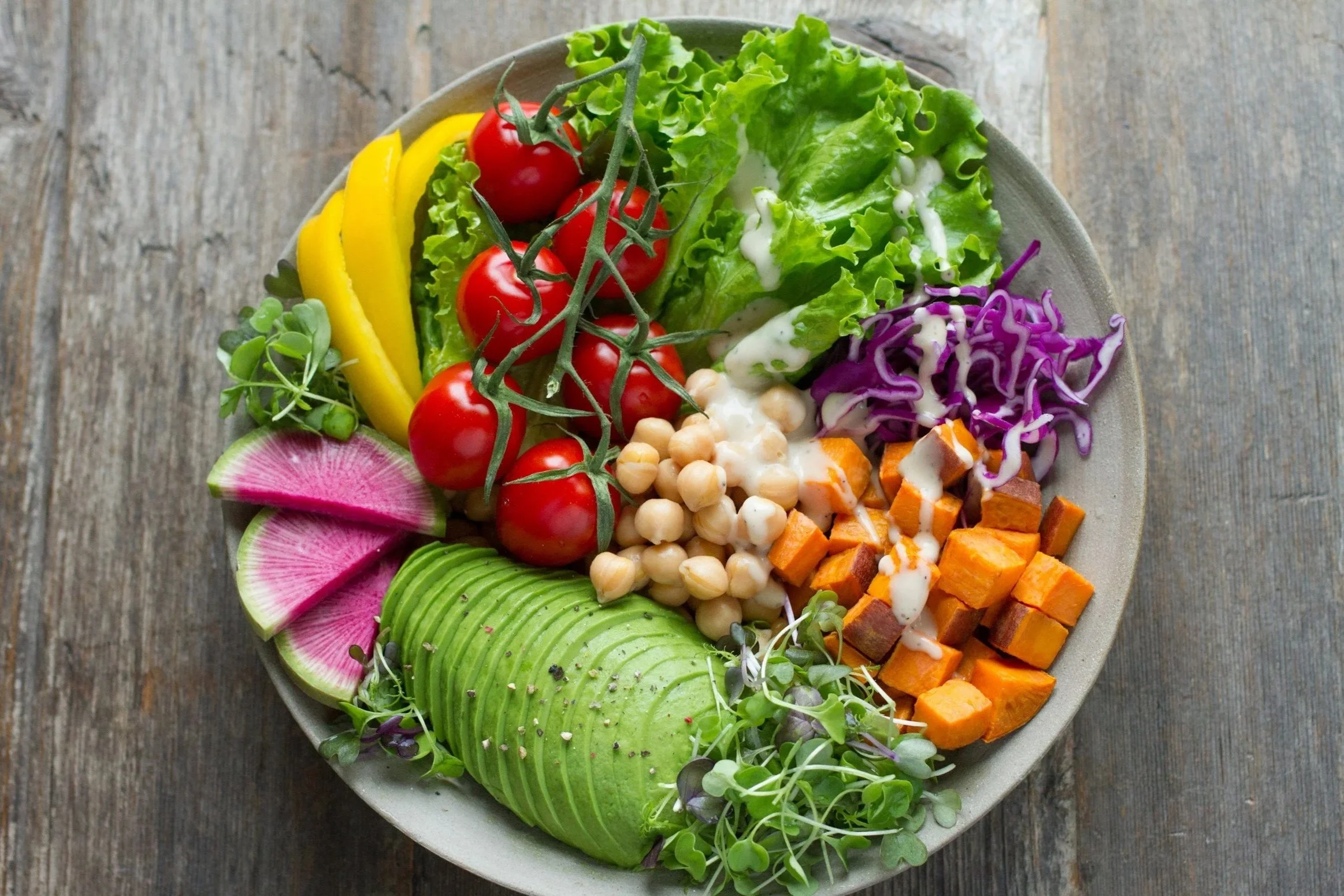 Assorted fresh vegetable salad on a round plate, including cherry tomatoes, sliced yellow bell peppers, radishes, avocado slices, green lettuce, purple cabbage, chickpeas, diced sweet potatoes, microgreens, and a drizzle of dressing, on a rustic wooden table.