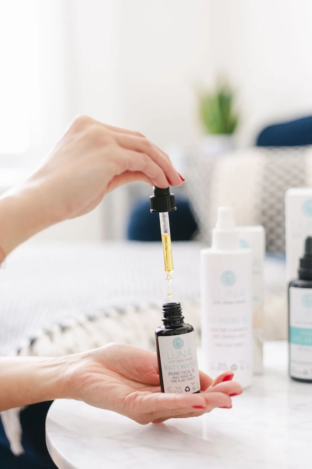 Person holding a bottle of Luna Organics skincare oil with a dropper, dispensing a yellow liquid into the bottle, on a white surface with other skincare products in the background.