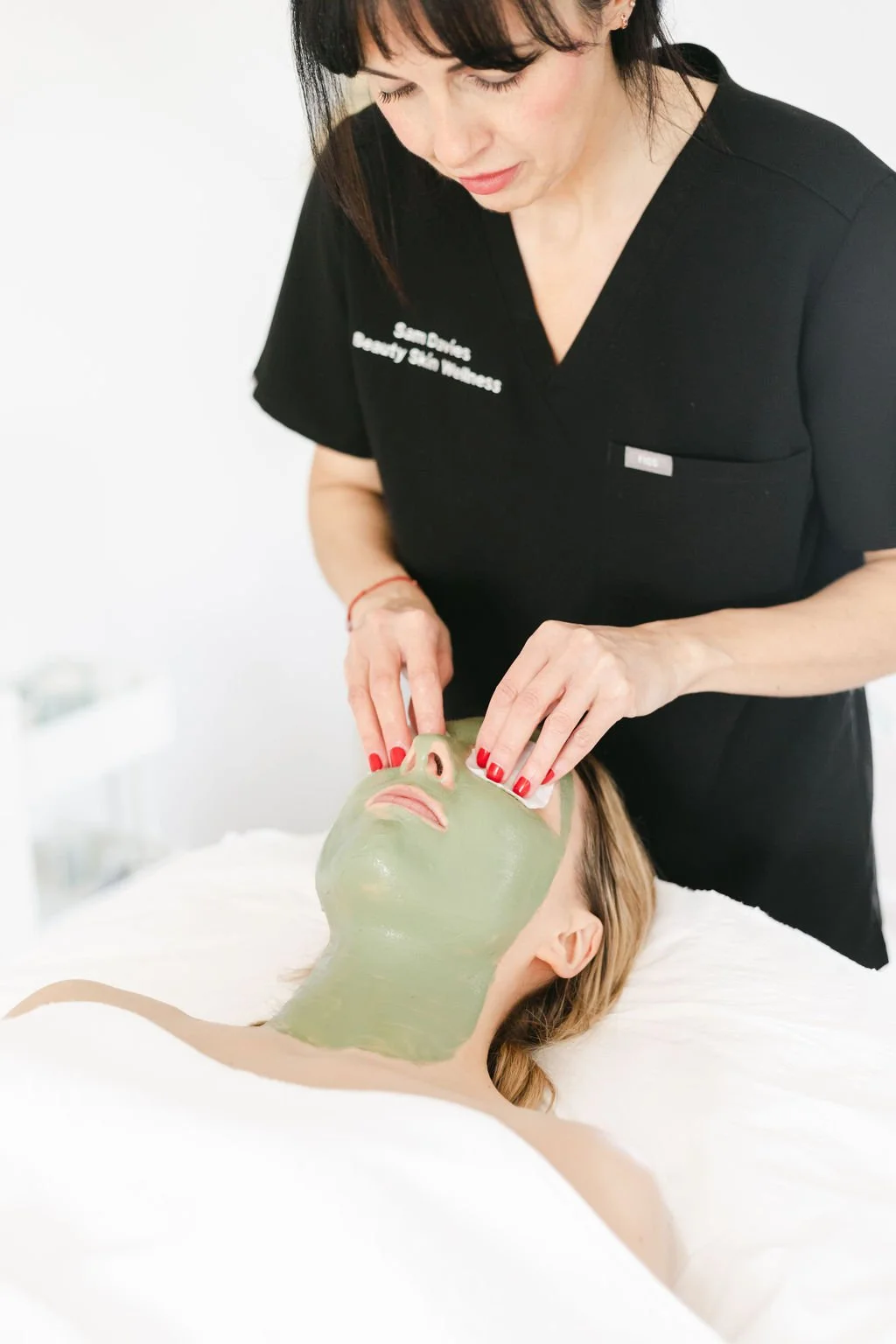 A woman is performing a facial treatment on another woman lying on a treatment bed, with her face covered in a green facial mask.