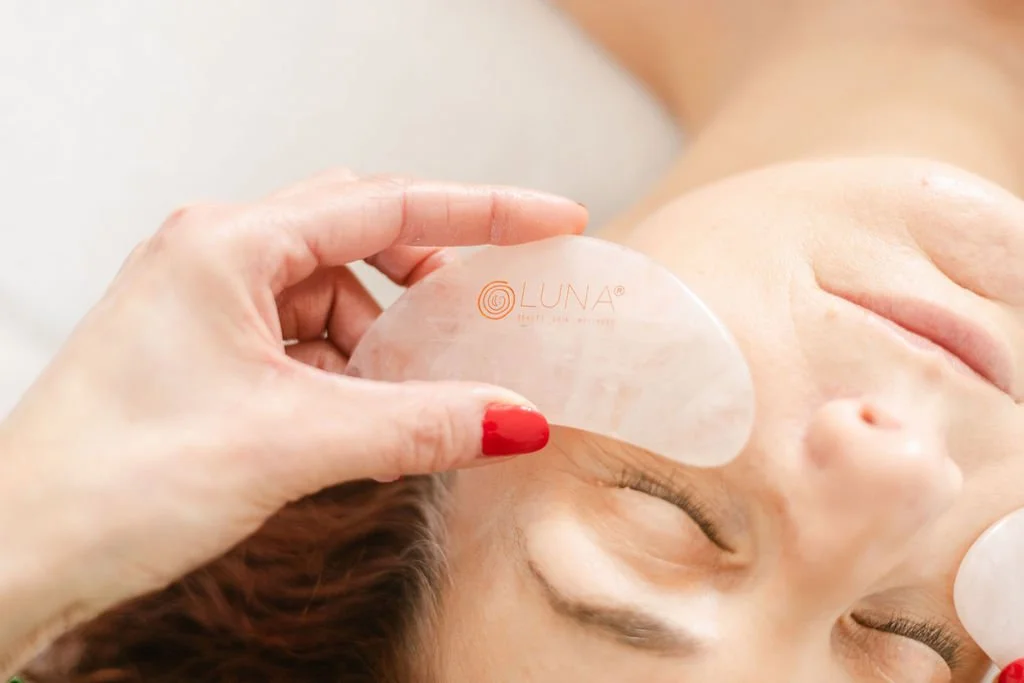 A person with brown hair receiving a facial treatment with a Gua Sha tool on the forehead.
