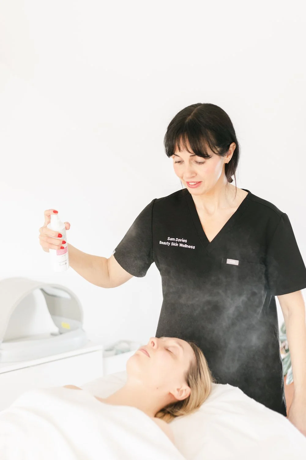 A woman is spraying a mist over a woman lying on a treatment bed in a spa or skincare clinic.