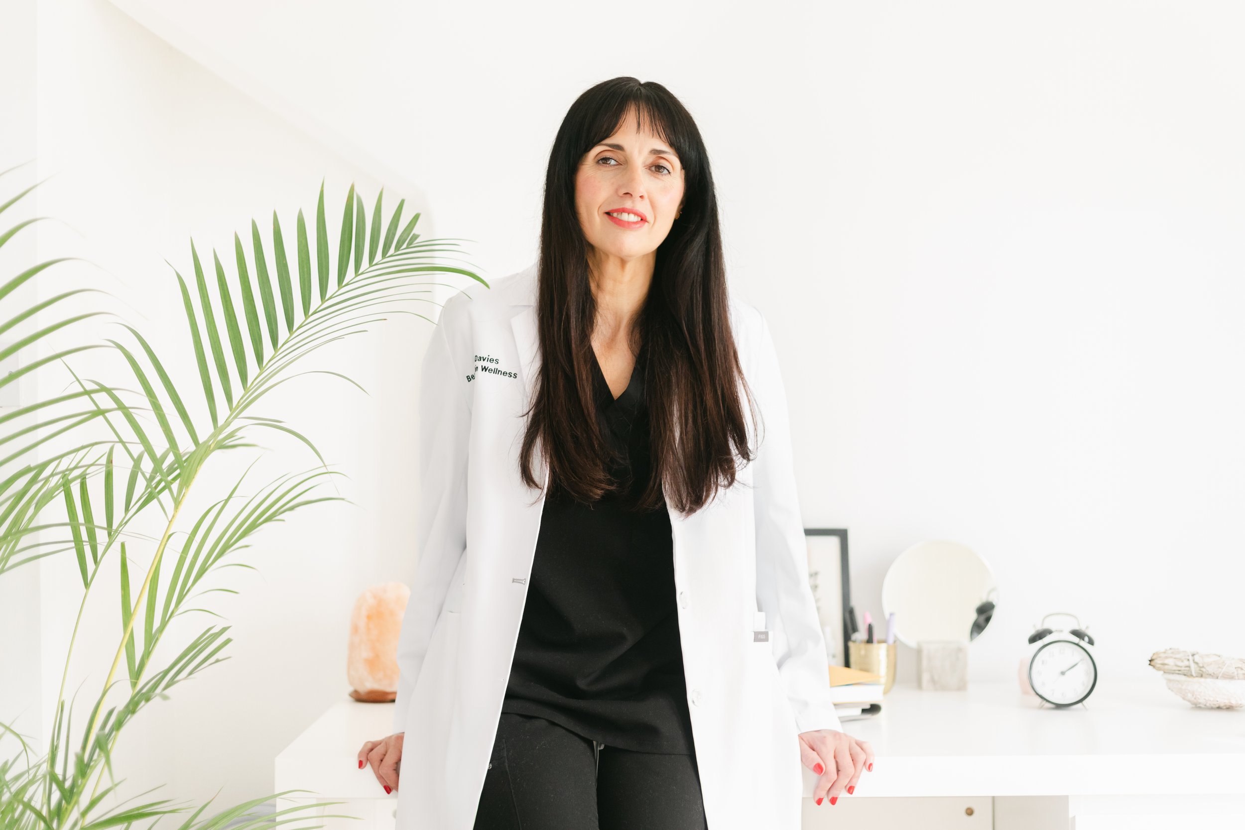 A woman with long dark hair and a white lab coat standing in a bright, minimalistic room with a white desk, a large green plant, and various small items including a salt lamp, framed picture, clock, and mirror.