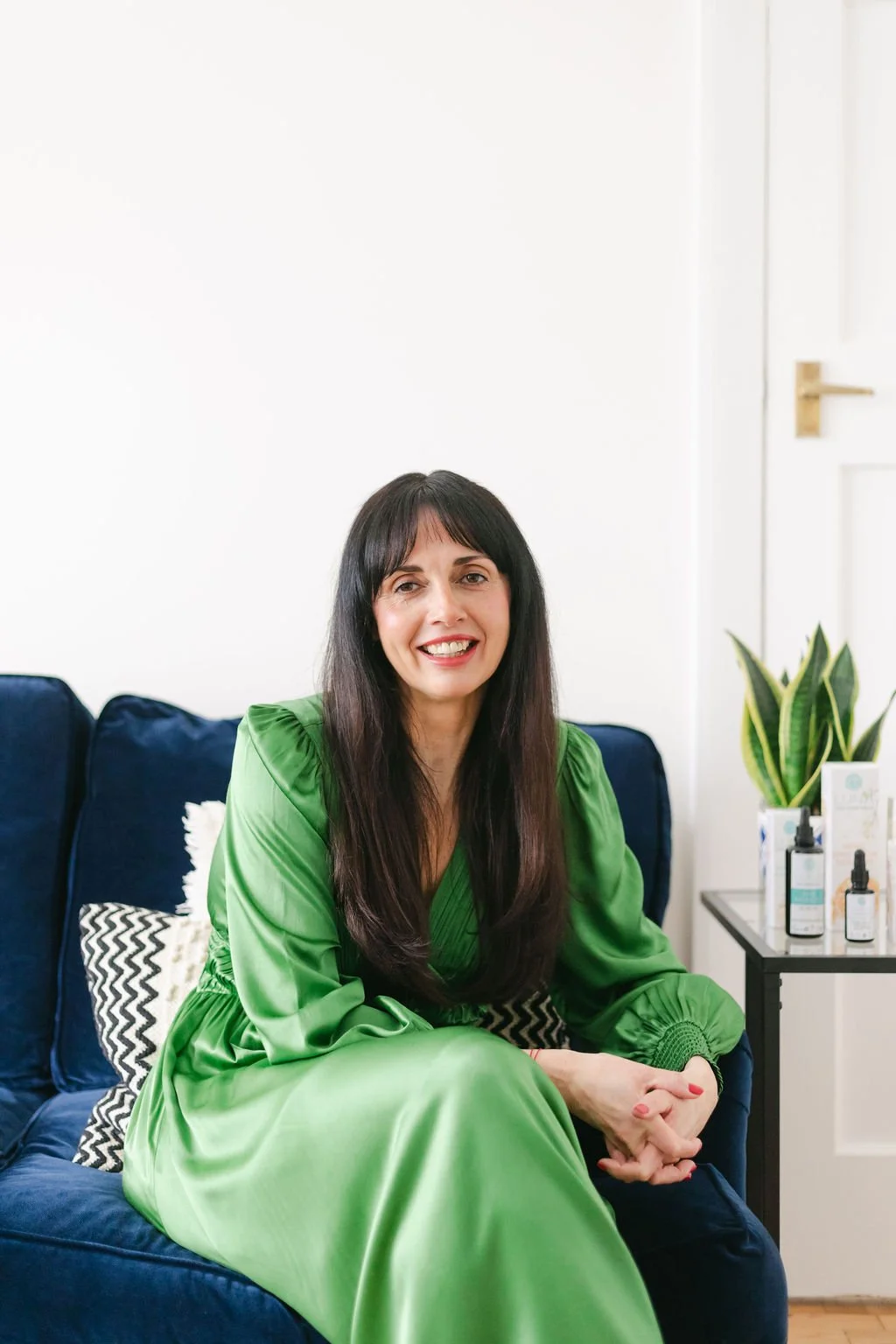 A woman with long dark hair and a bright smile sits on a navy sofa, wearing a green satin dress. In the background, there are bottles of skincare products on a side table and a plant.