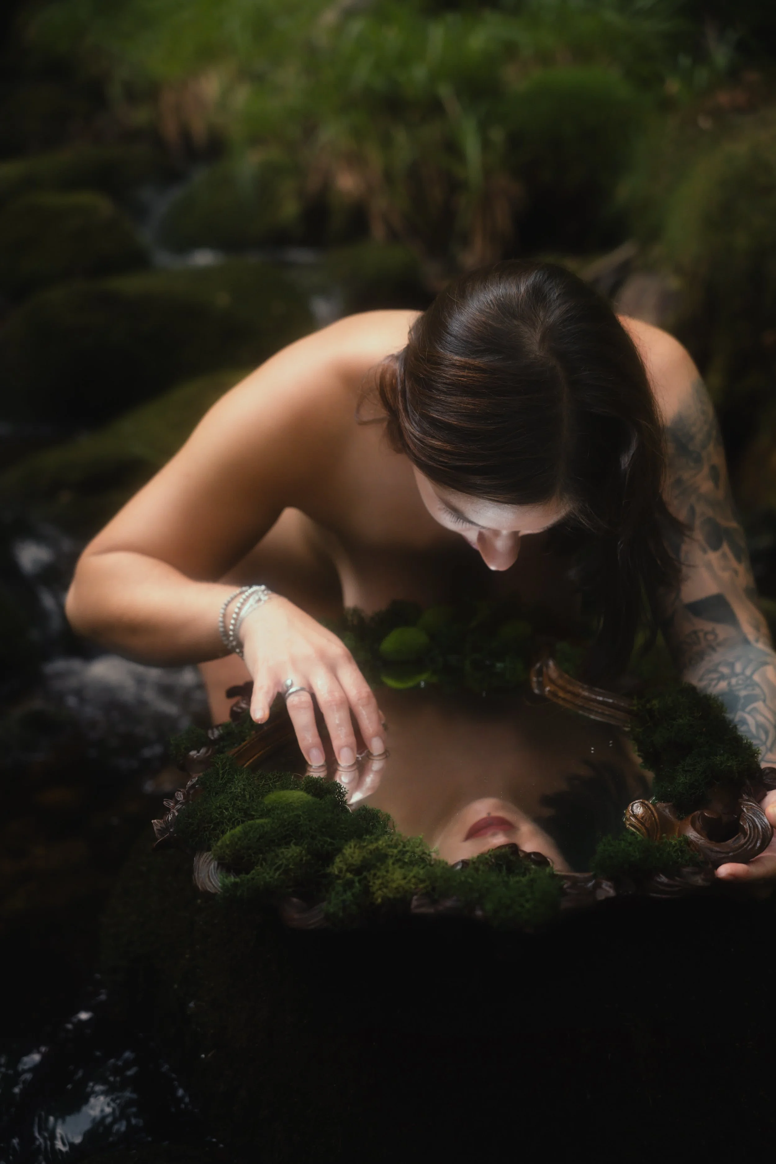 Photos nue artistique - Femme nue avec tatouages, penchée sur un miroir ancien entouré de mousse, dans un environnement naturel de forêt.