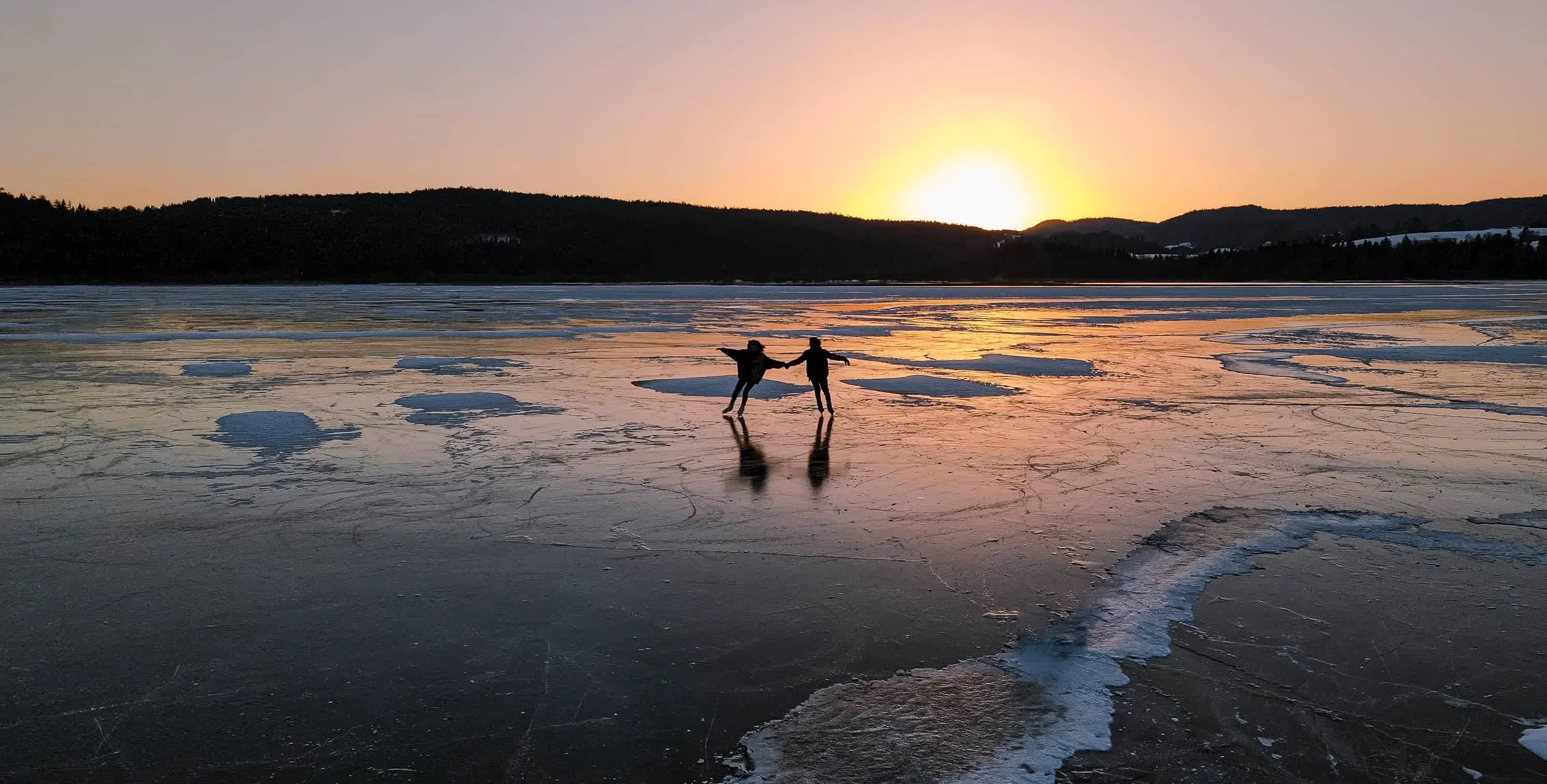 Deux personnes qui dansent sur un lac gelé au coucher du soleil, avec des montagnes en arrière-plan et des fragments de glace sur la surface du lac. Photo au drone 