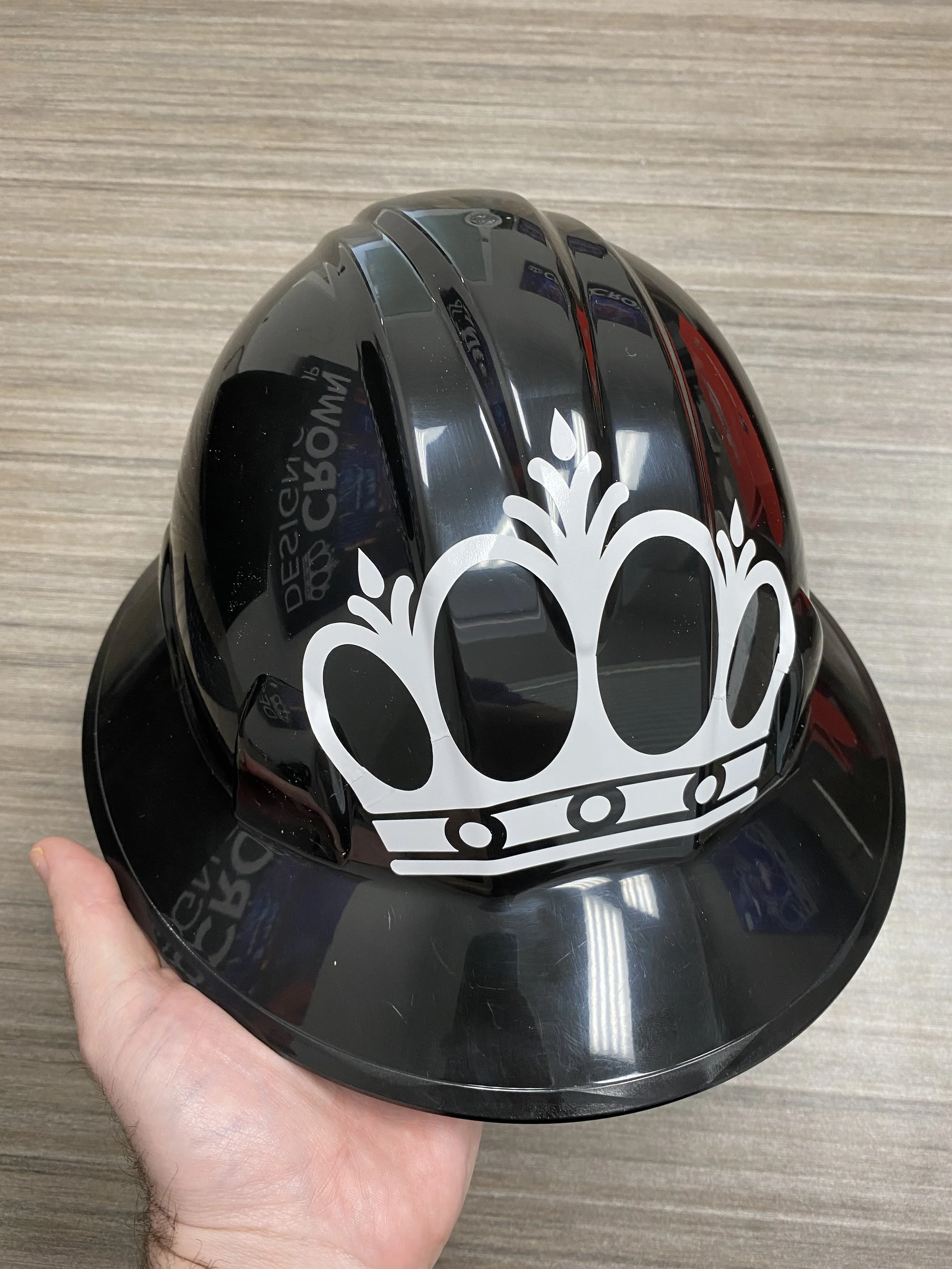 A black hard hat with a white crown decal held in a person's hand against a wooden background.