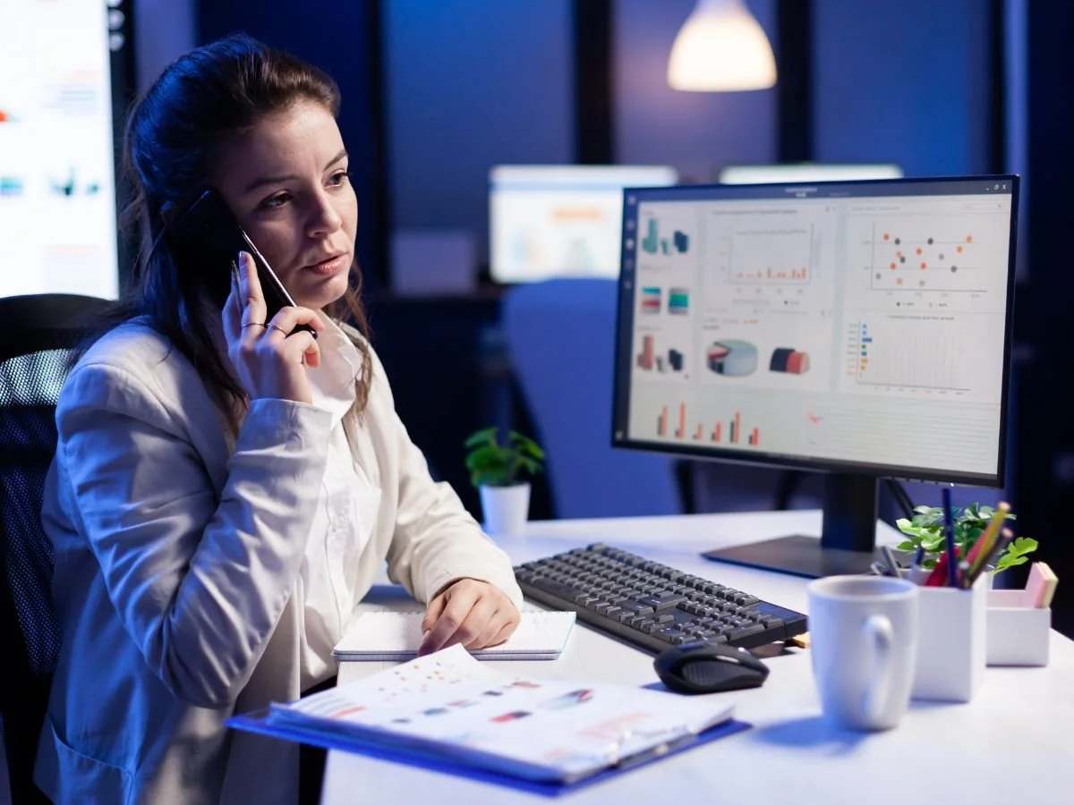 A woman in a white shirt sitting at a desk, talking on the phone, with a computer monitor displaying analytics and charts, a notebook, a cup, and office supplies.