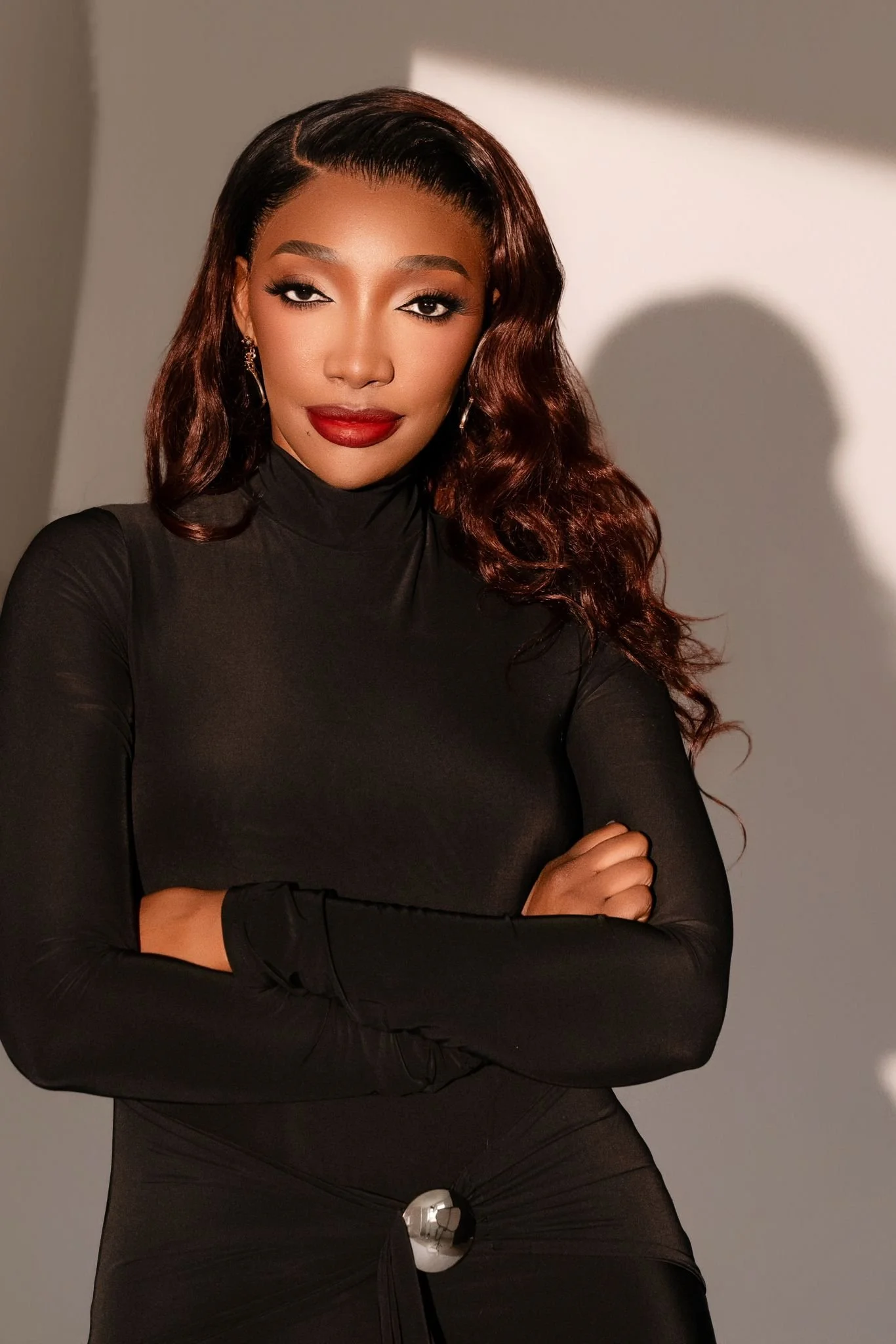 A woman with long, wavy brown hair, wearing a black turtleneck top, cross arms, standing against a plain light gray background with a shadow cast on the wall.
