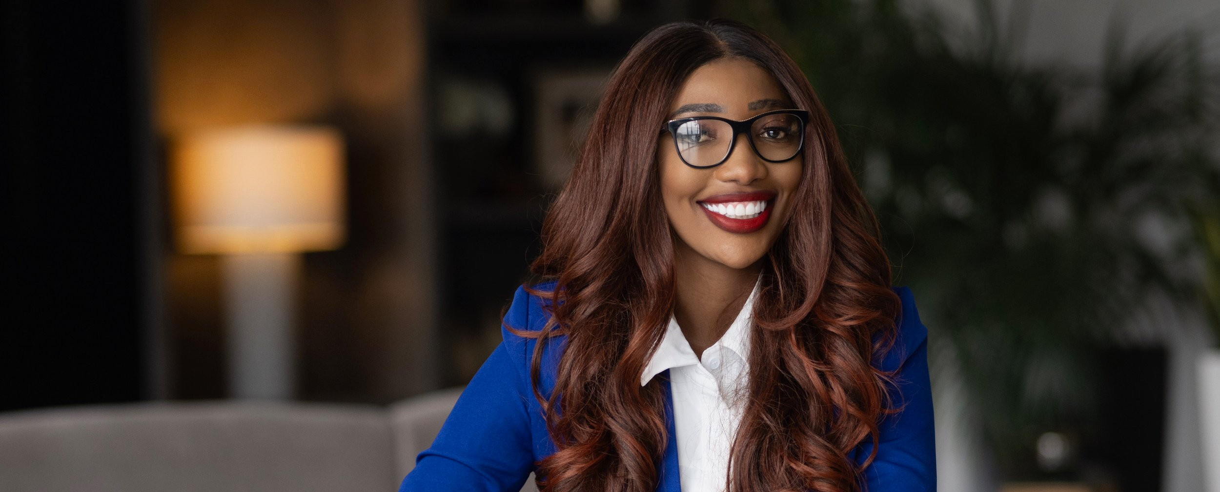 A woman with long wavy auburn hair, wearing glasses, a white shirt, and a blue blazer, smiling while sitting in a cozy room with warm lighting and a blurred background including a lamp and plants.