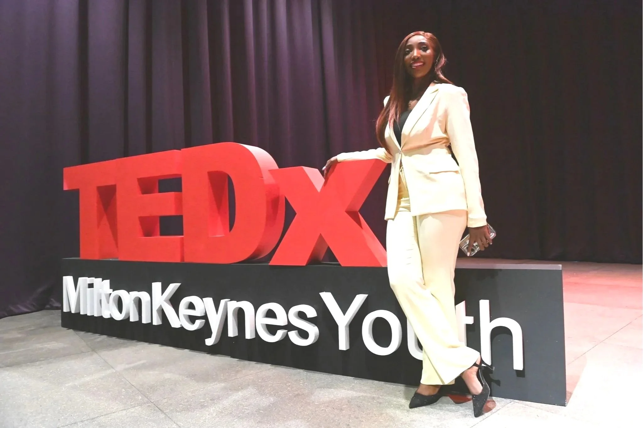 A woman in a cream-colored suit and black high heels standing next to a large red TEDx sign with 'MiltonKeynesYOUTH' written below, on a stage with black curtains in the background.