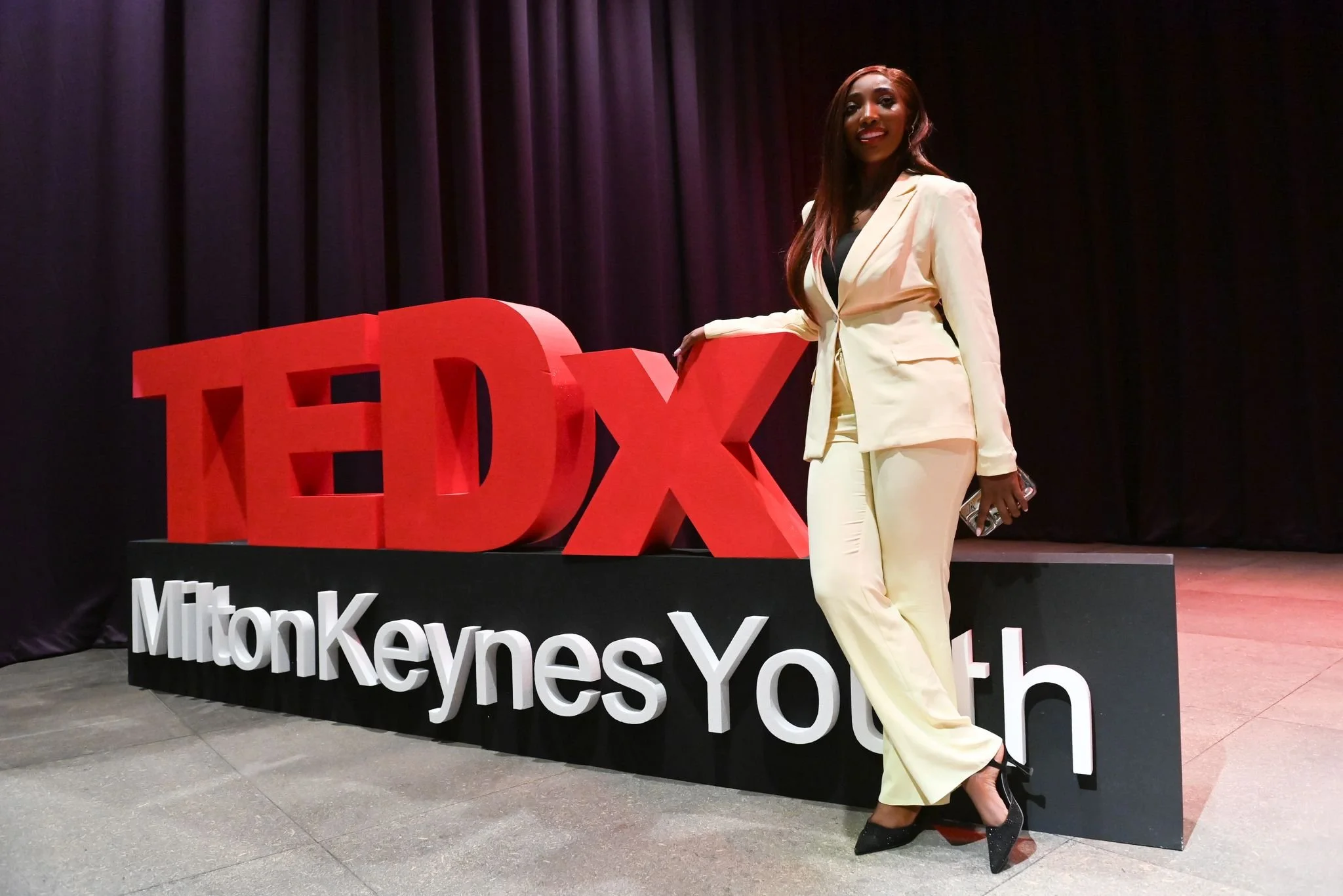 A woman in a light-colored suit posing next to a large red TEDx sign at a youth event in Milton Keynes.