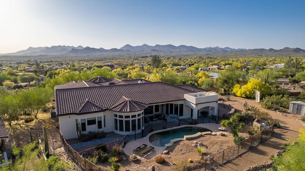 Aerial view of a suburban house with a tiled roof and backyard pool, surrounded by desert landscape and mountains in the background under a clear sky.