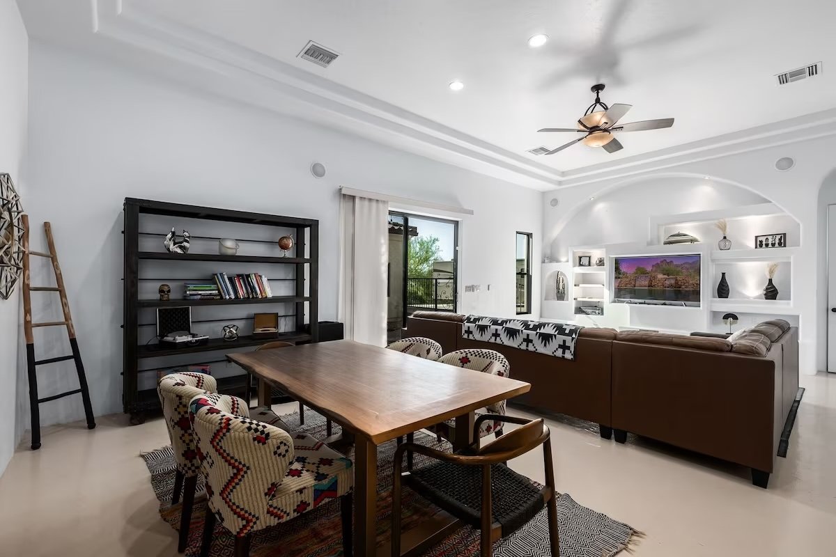 Modern living and dining area with a wooden table, patterned chairs, brown leather sectional sofa, built-in white shelves, flat-screen TV, bookshelf with decor, ladder, and ceiling fan.