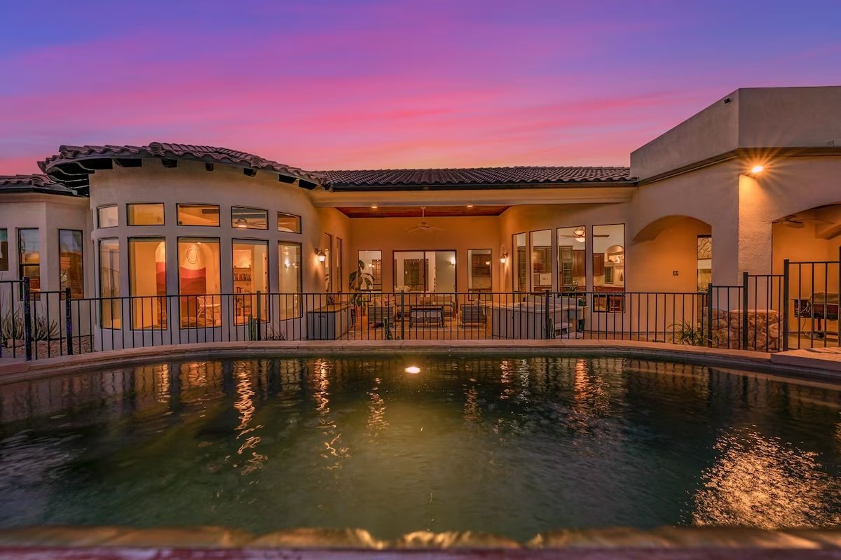 Modern house with large windows overlooking a swimming pool at sunset, featuring a fenced patio and warm exterior lighting.