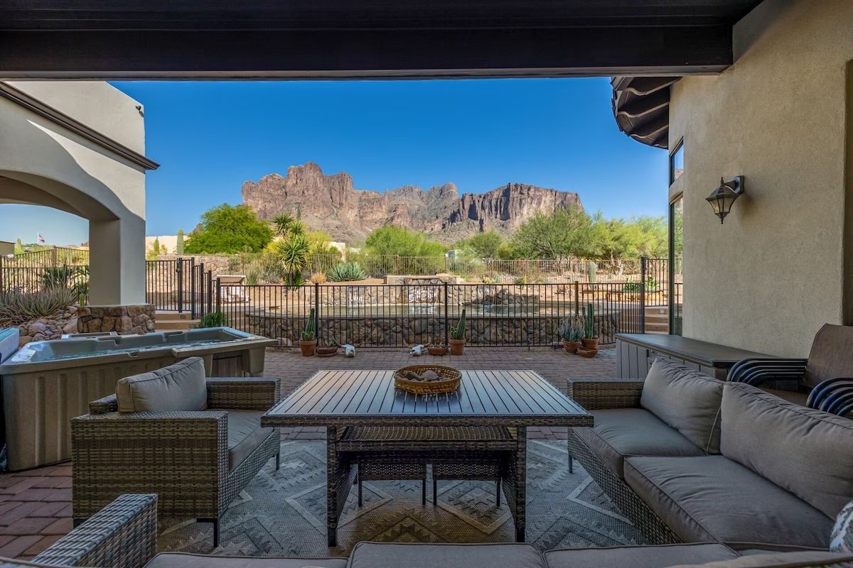Outdoor patio with cushioned furniture overlooking a fenced backyard and desert landscape with mountains in the background.