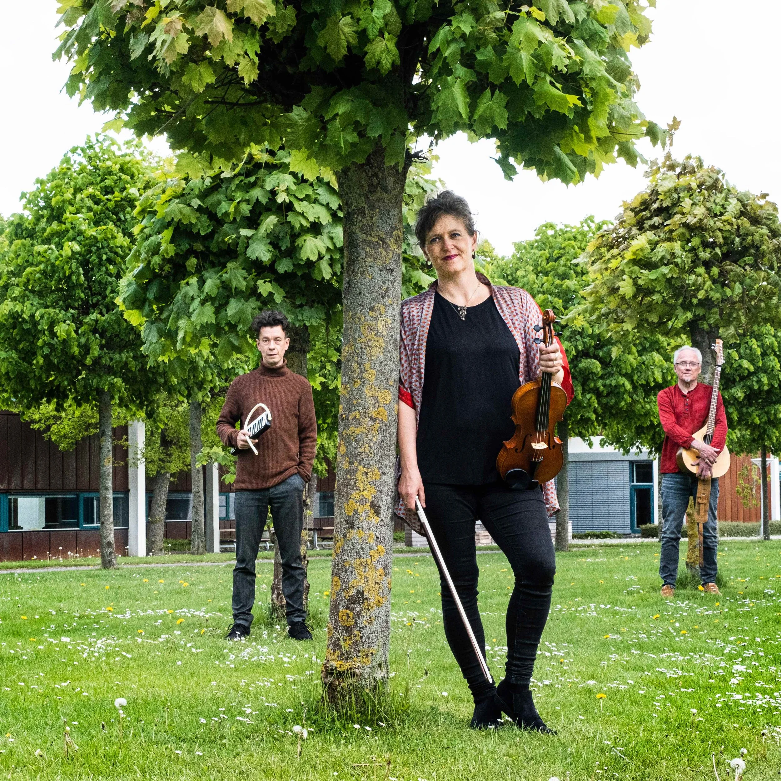 Press photo of Trio Mio. Three musicians standing outdoors on a grassy area with trees, holding their instruments. The woman in the foreground has a violin, the man on the left has a melodica, and the man on the right has a bouzouki.