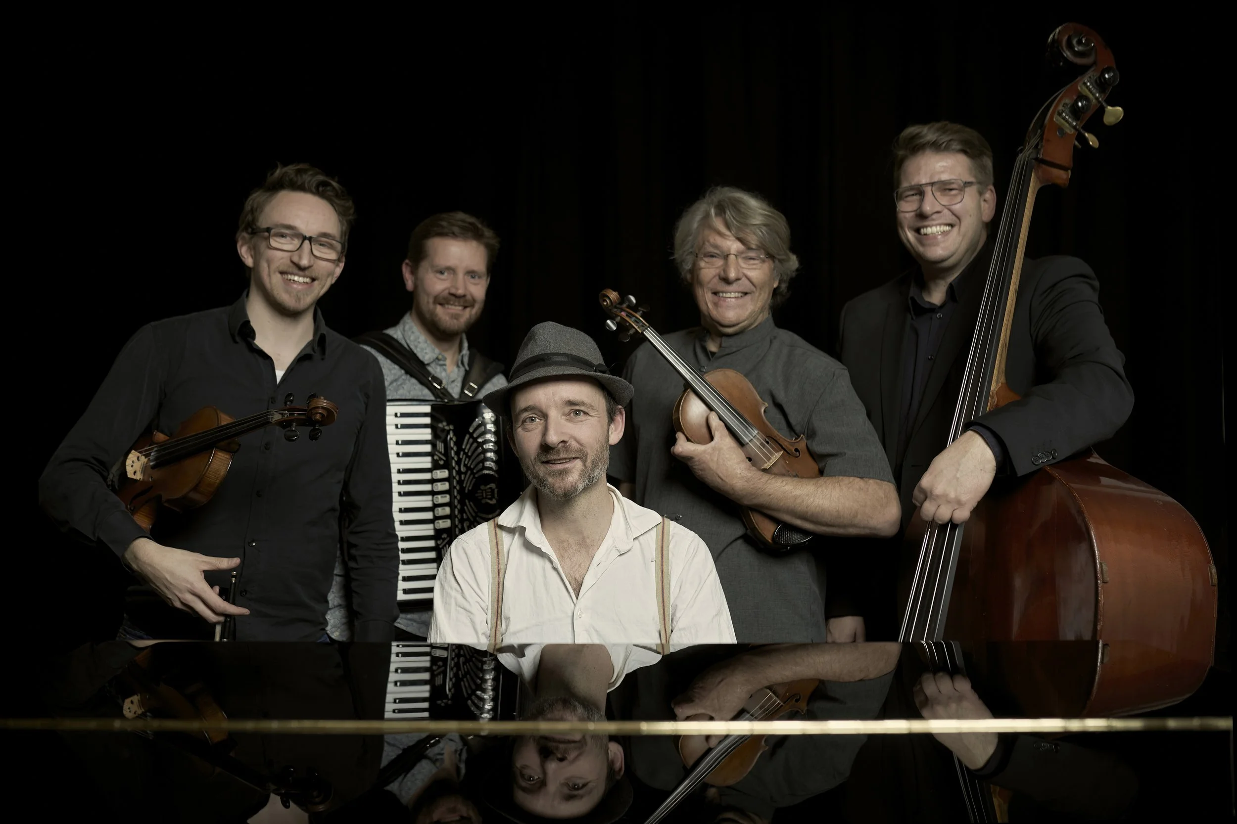 Press photo of Fionia Folk Brothers. Group of five male musicians holding musical instruments, including a violin, accordion, double bass, and a piano, smiling in a studio with a black background.