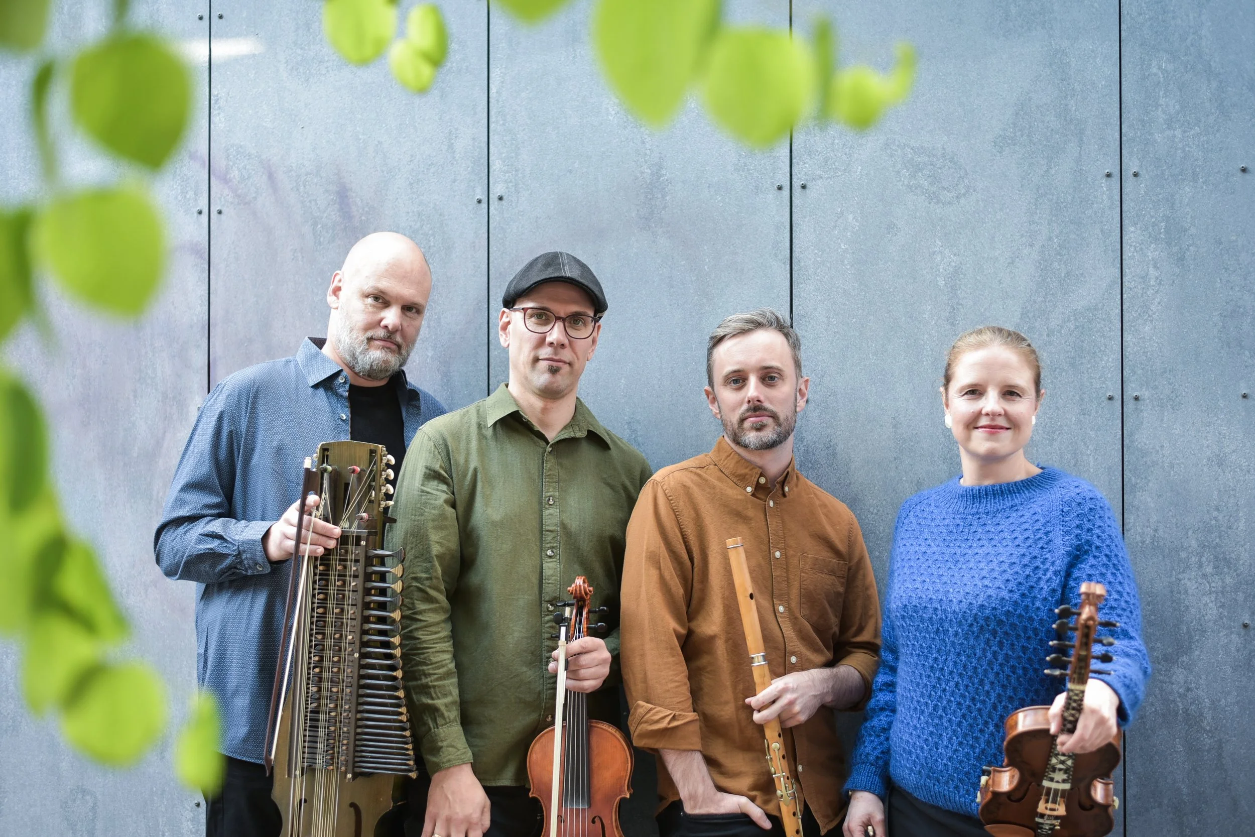 Press photo of Purppuri. Four musicians stand against a blue-gray wall, each holding a string instrument including violins, a viola, and a violin, with one woman wearing a blue sweater and the three men dressed in shirts.
