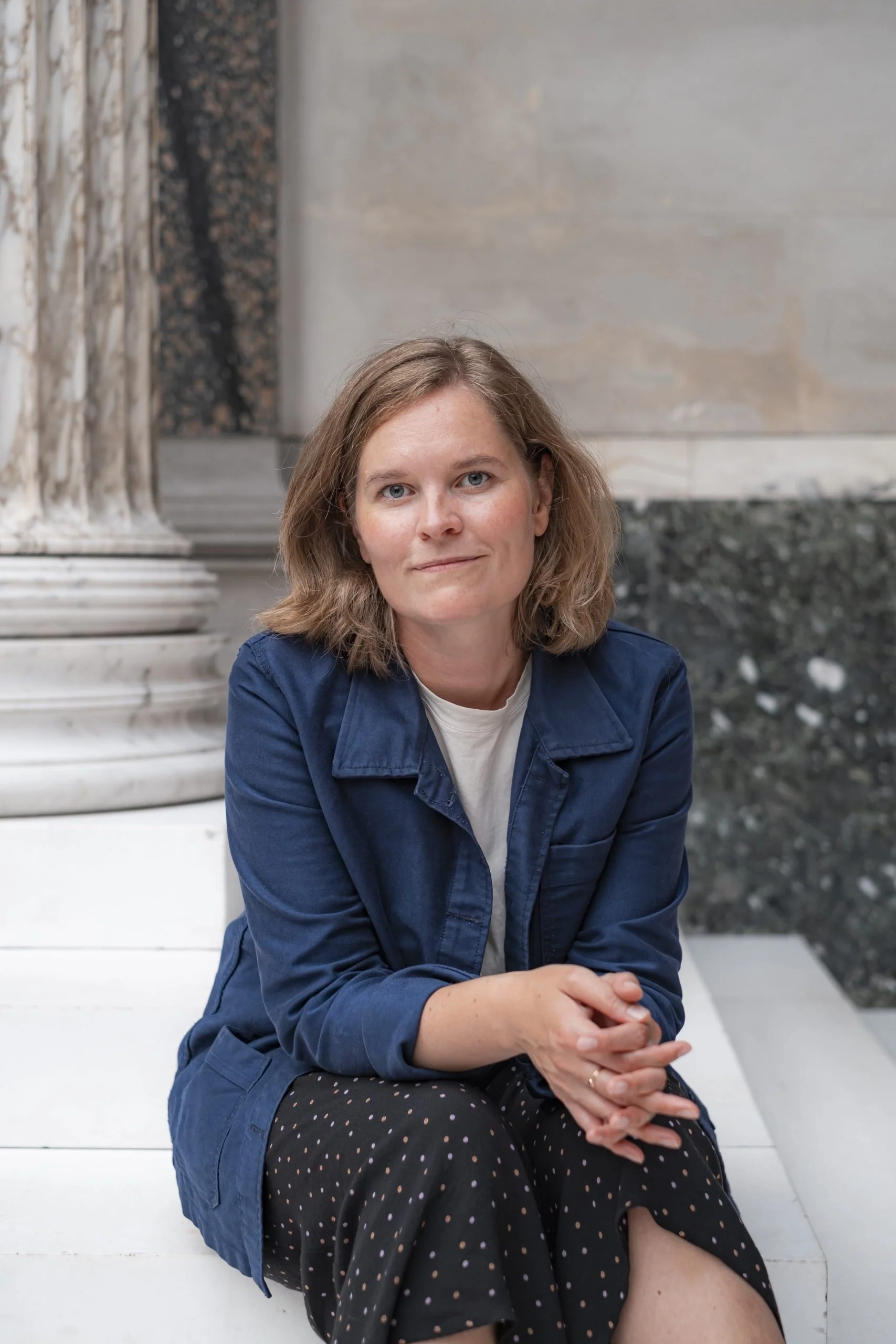 Portrait of a moderator Benedikte Granvig. She has short brown hair, wearing a navy blue jacket over a white shirt, sitting on white steps inside a building with marble pillars and walls.