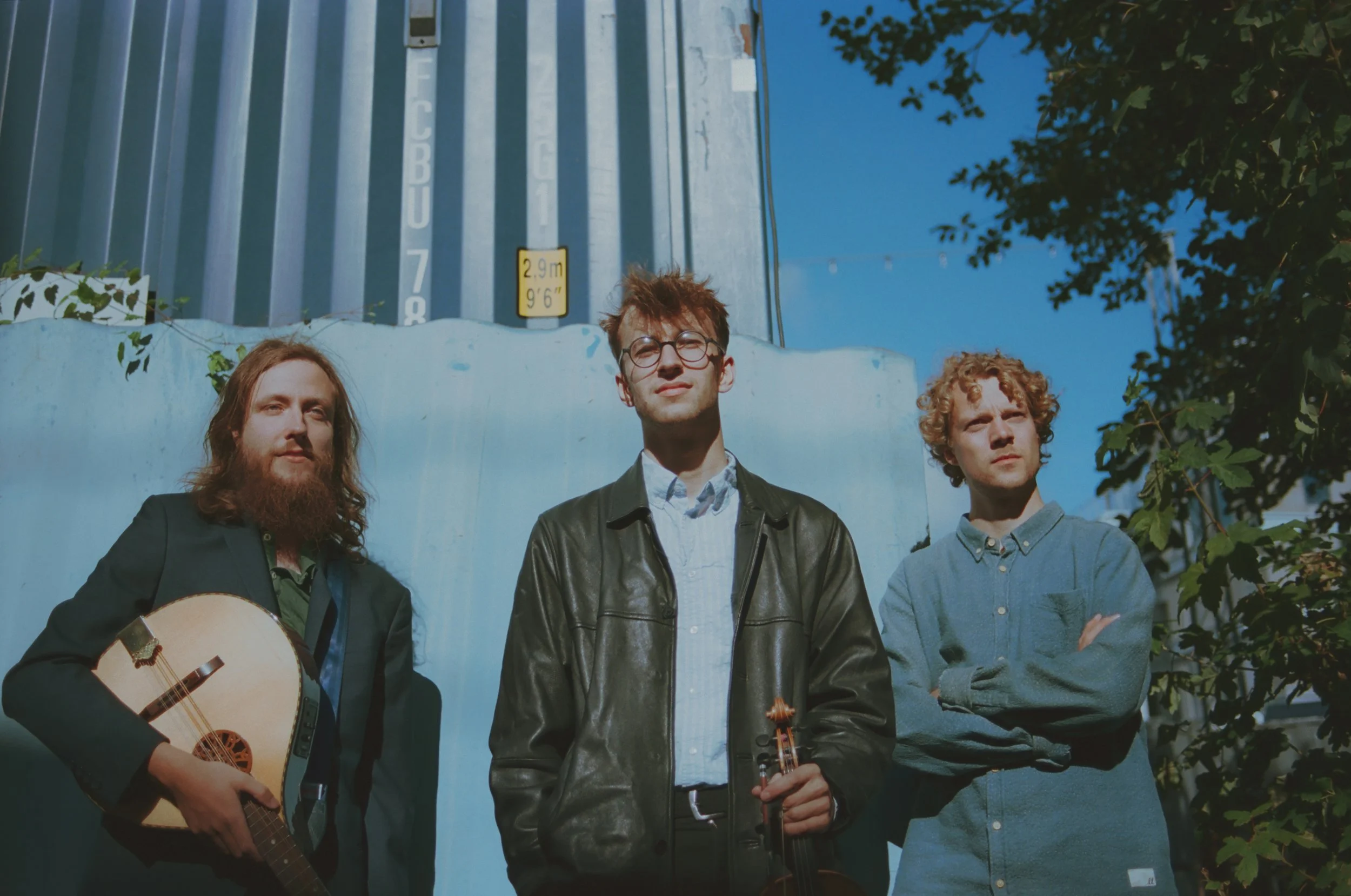 Press photo of Carol Supreme. Three young men standing outdoors under a blue sky, with a large shipping container in the background. One is holding a bouzouki, another a violin, and the third has arms crossed.