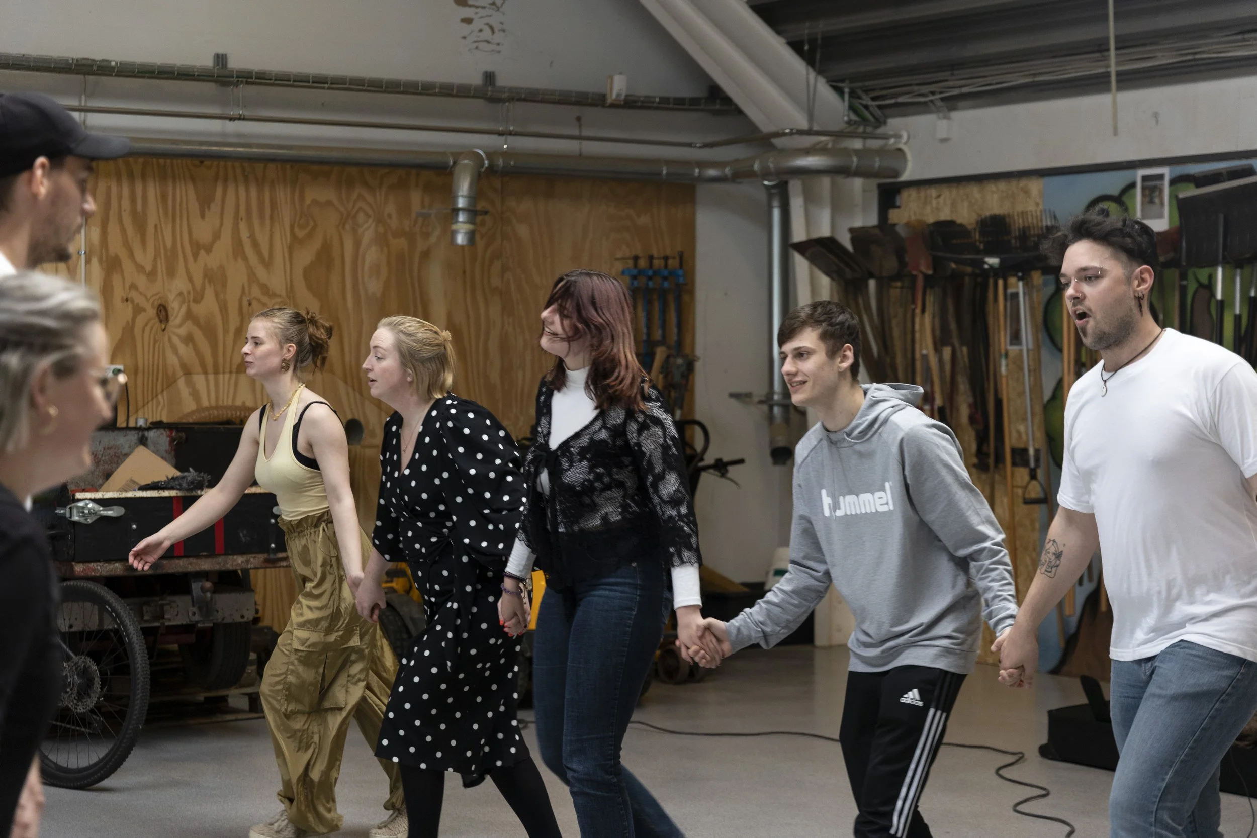 Group of people holding hands in a circle, practicing dance in a studio with storage racks and tools in the background.