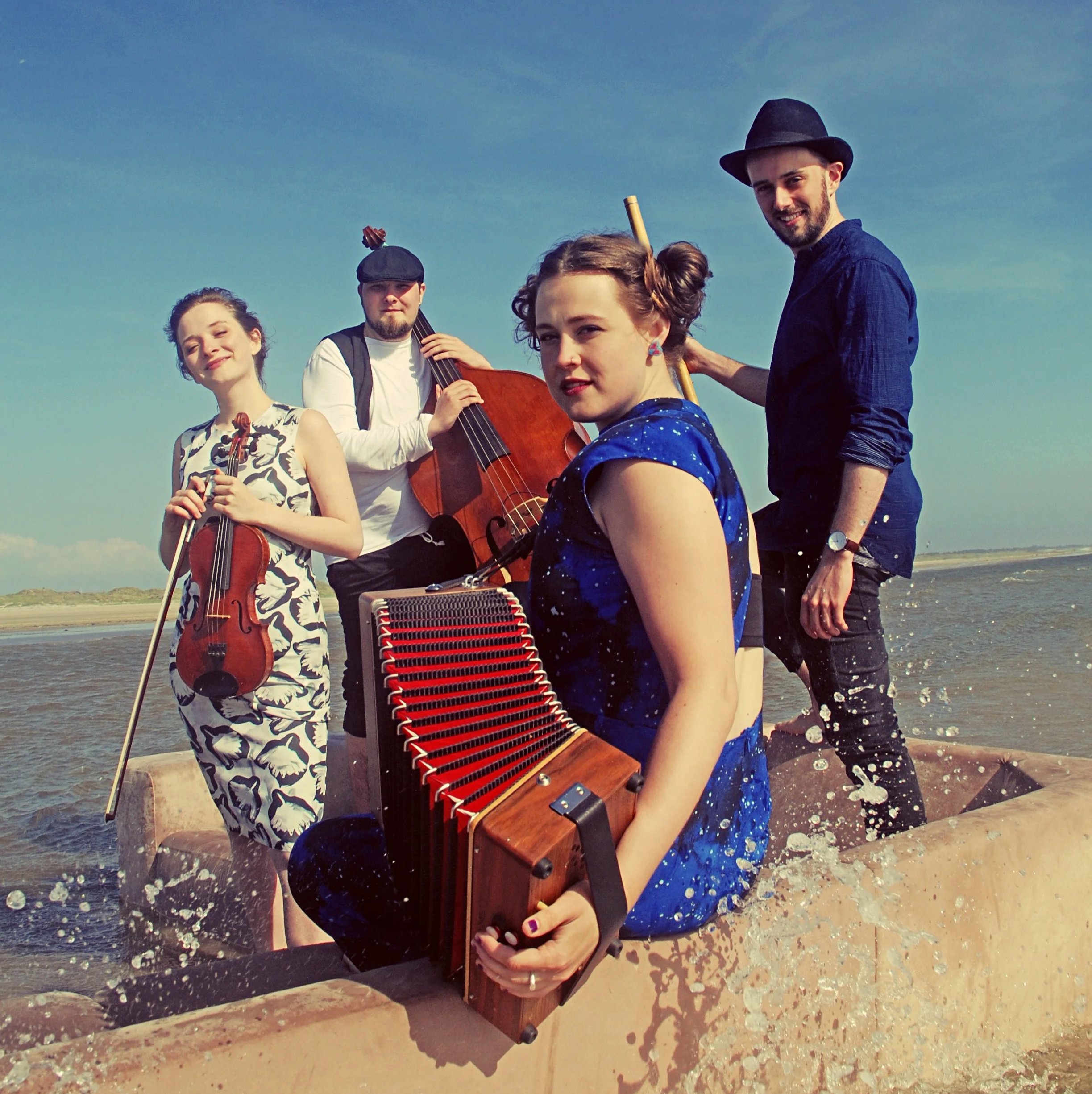 Press photo of Floating Sofa Quartet. Group of four musicians on a boat in the water, holding instruments including a violin, double bass, accordion, and a flute, with a blue sky in the background.