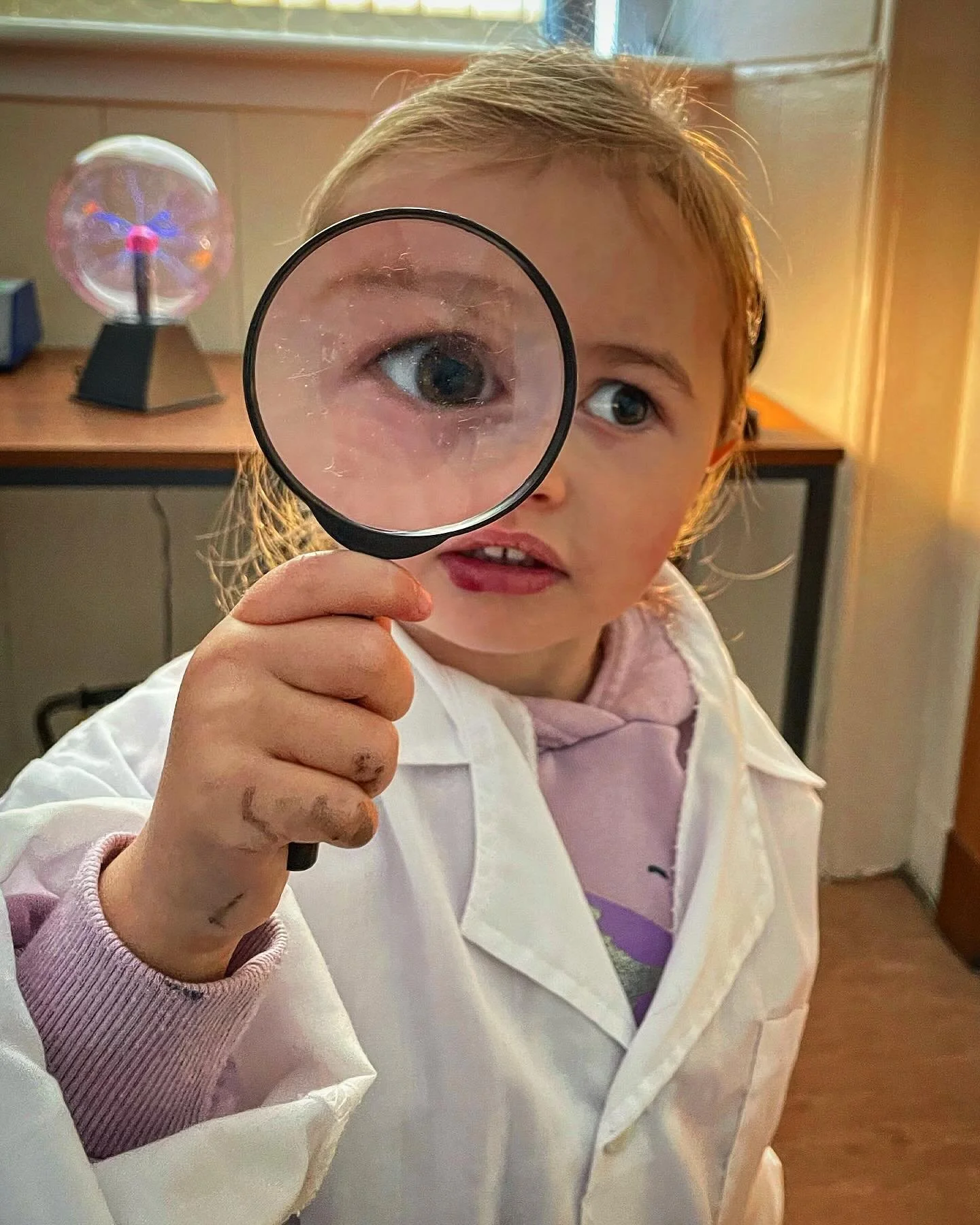 Child holding a magnifying glass with an eye visible through it, wearing a white lab coat and a pink hoodie, in a room with a small Tesla coil in the background.