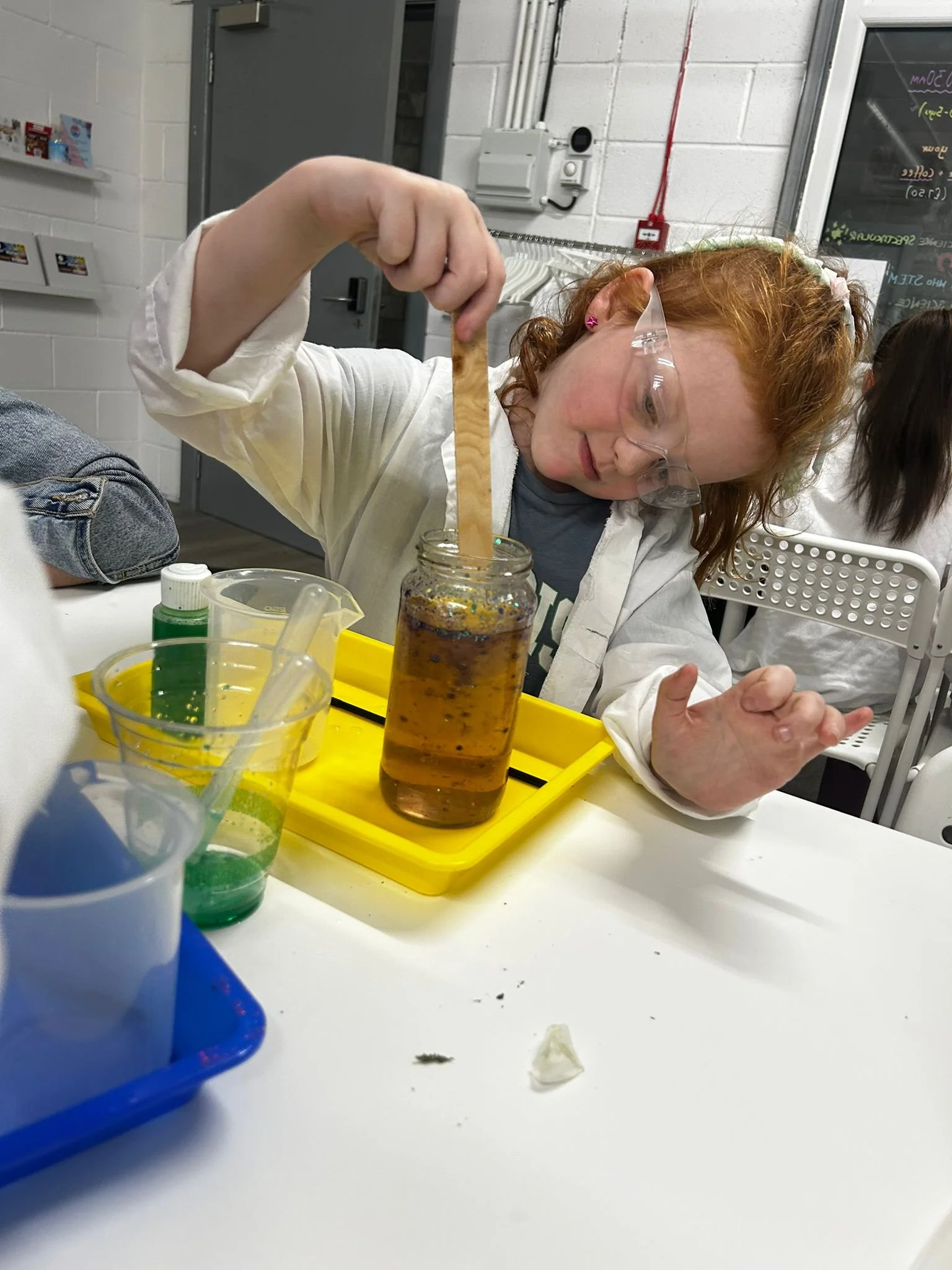 A girl with red hair, wearing safety glasses, is adding a liquid to a jar containing a bubbling, dark-colored mixture in a science classroom.