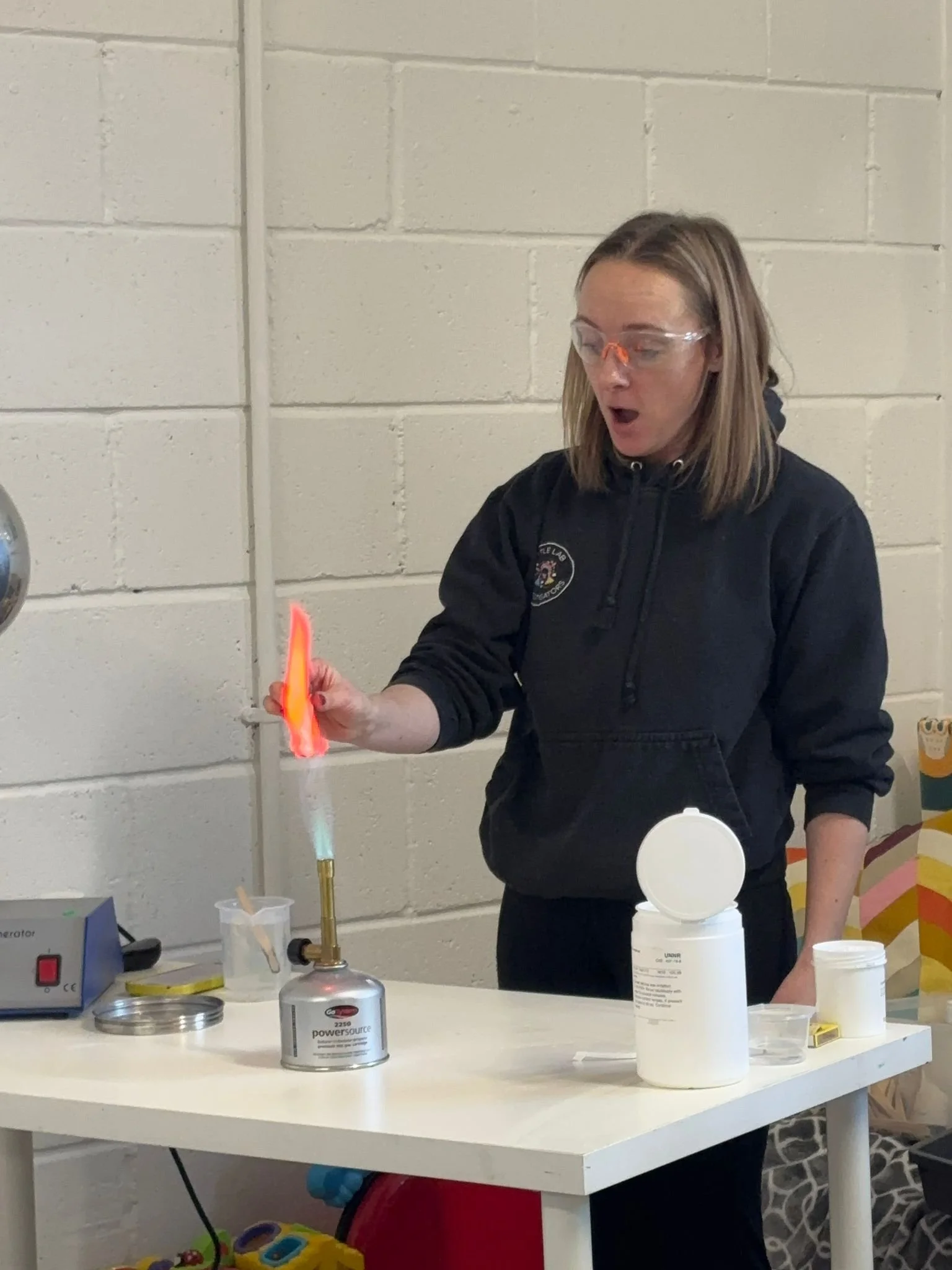 A woman in safety glasses and a black hoodie is holding a piece of orange material over a Bunsen burner flame, which is producing a blue flame and a small flame. She appears to be conducting a science experiment in a laboratory or classroom setting.