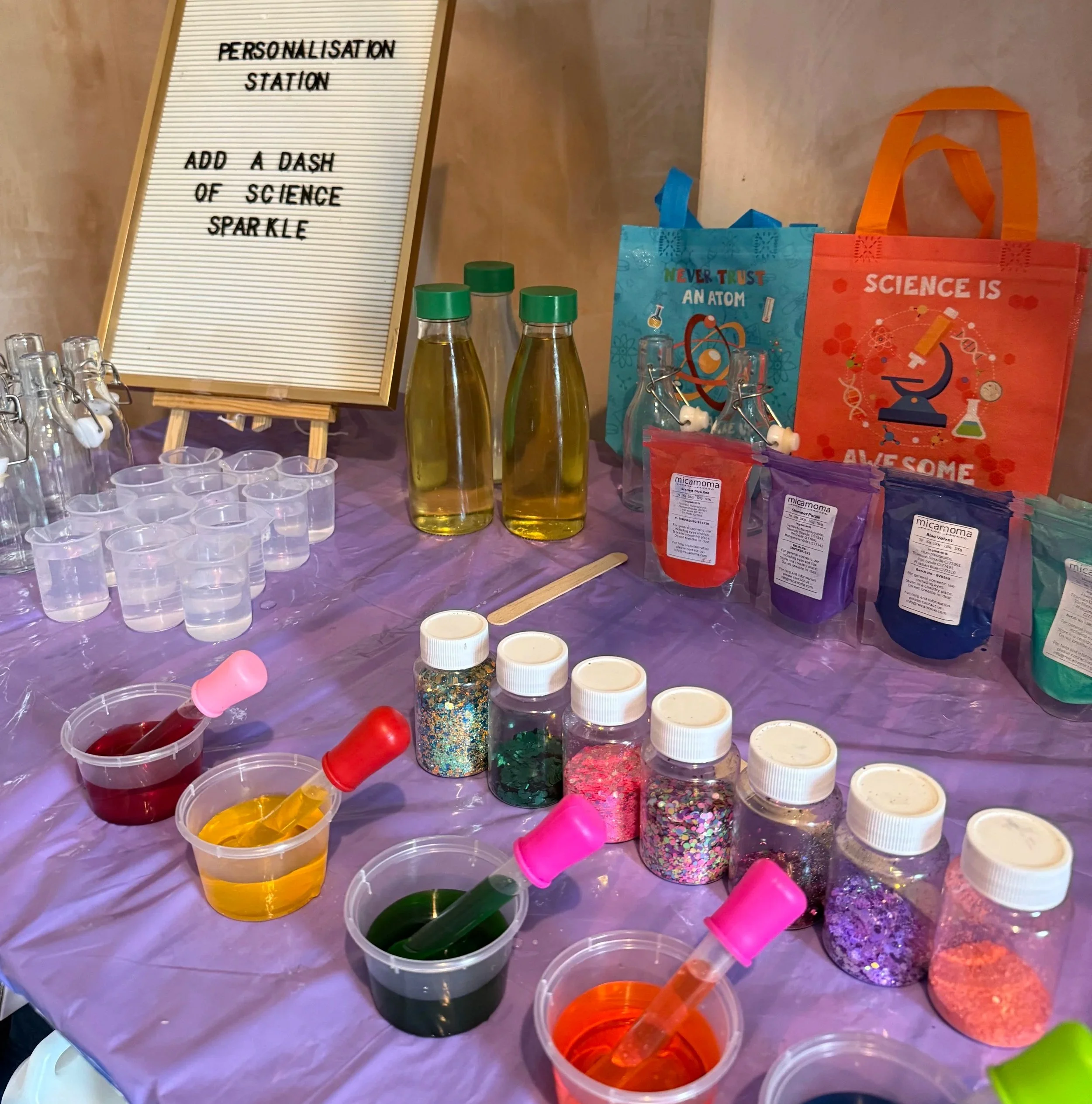 A science-themed personalisation station with bottles of liquid, small cups, jars of colorful glitter, and bags of coloring powder on a purple tablecloth. There is a sign that reads 'PERSONALISATION STATION ADD A DASH OF SCIENCE SPARKLE' and colorful bags with science-themed designs.
