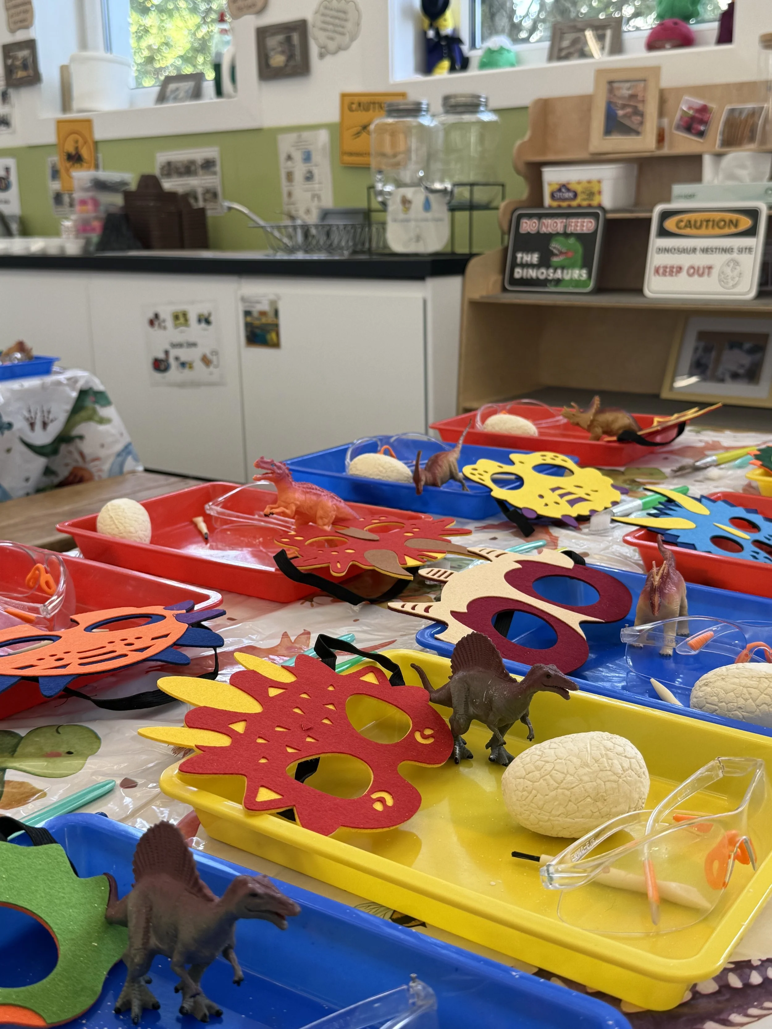 Play food and toy dinosaurs on colorful trays, with some eggs and safety goggles, in a classroom or craft room setting with signs and pictures in the background.