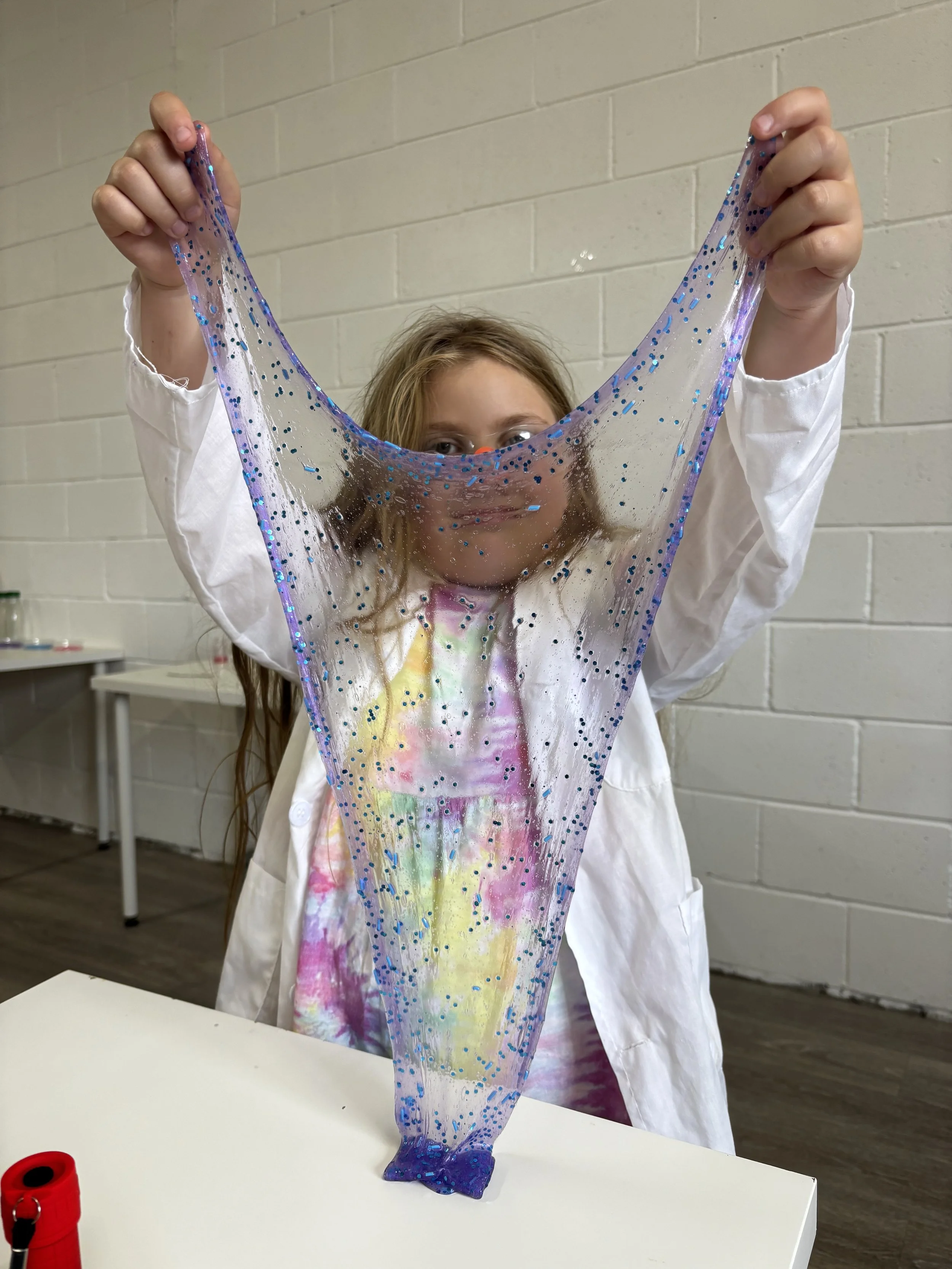 A young girl in a white lab coat holding up a large, transparent slime with blue glitter, stretching it in front of her face.