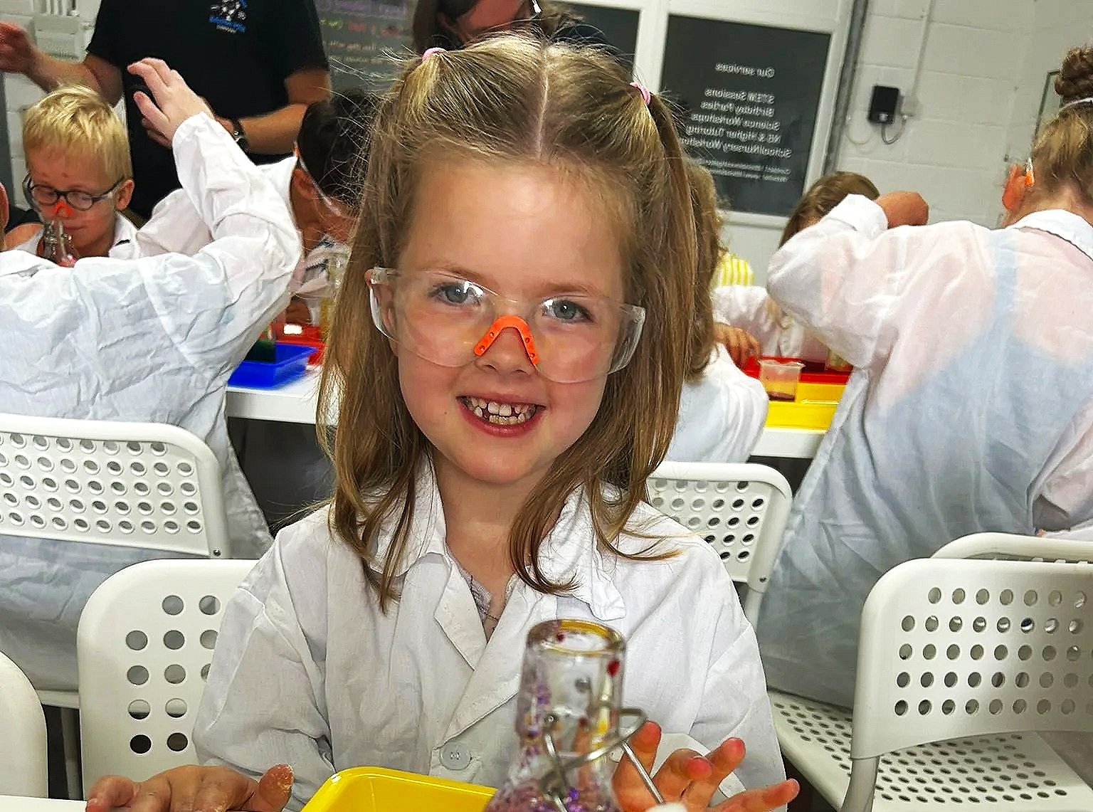 A young girl with red hair, wearing safety goggles and a white lab coat, smiling at the camera during a science experiment in a classroom, with other children working at tables in the background.
