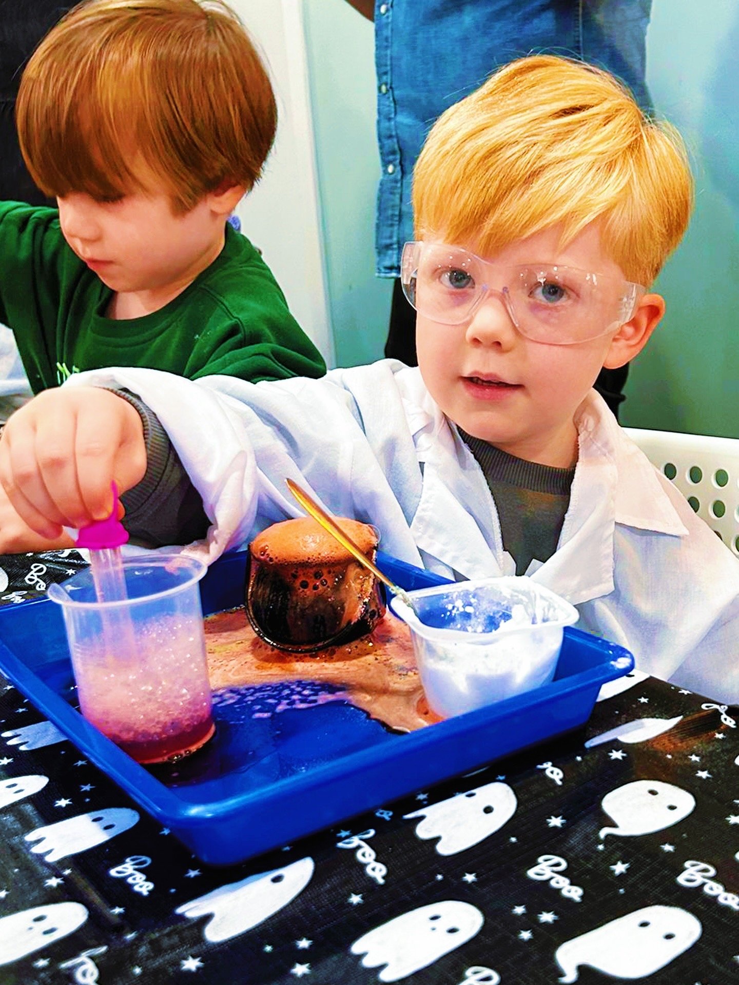 A young boy with red hair and safety glasses is engaged in a science experiment at a table. He is holding a pipette and working with colored liquids in a clear cup. There is a small container of foam or cream on the tray in front of him, and the tablecloth has a Halloween ghost design.