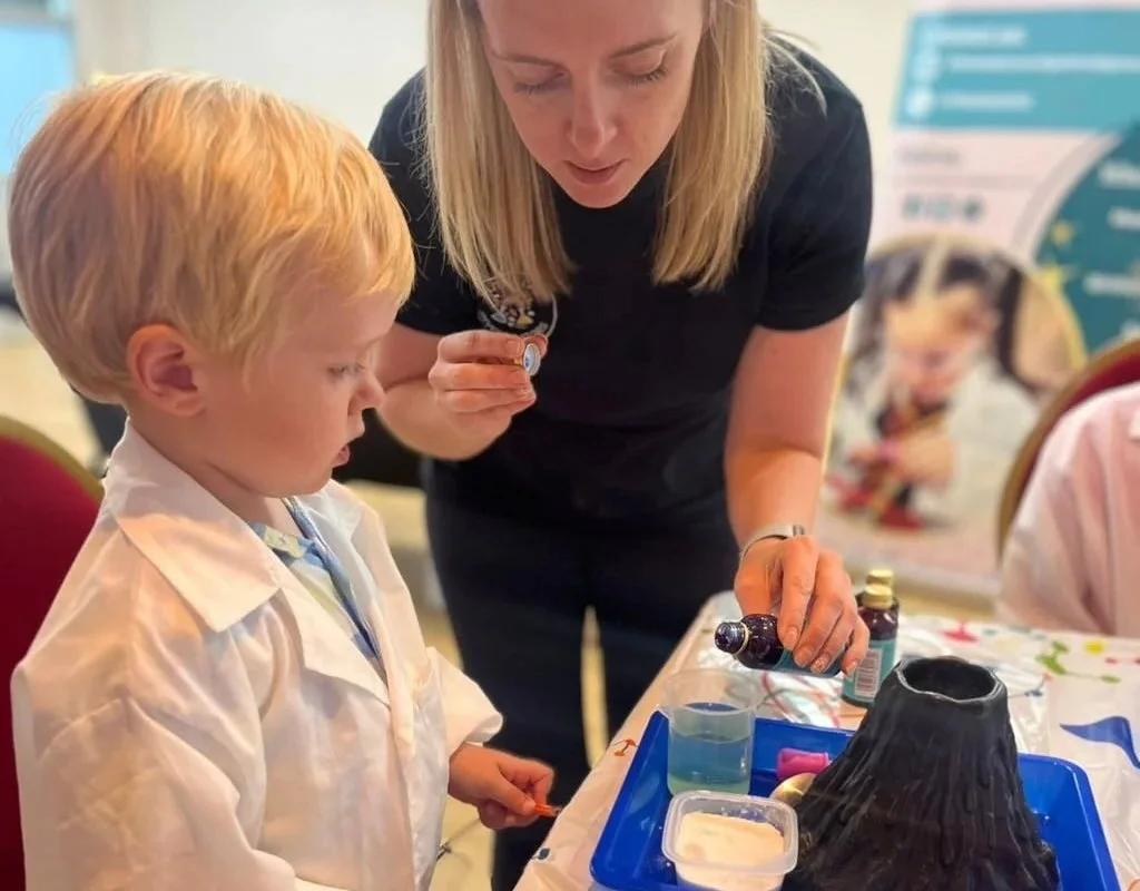 A woman with blonde hair helps a young boy with red hair blow bubbles at a science activity table. The table has items like a container with liquid, a funnel, and a volcano-shaped object. In the background, there is a poster with a photo of a child and some informational text.