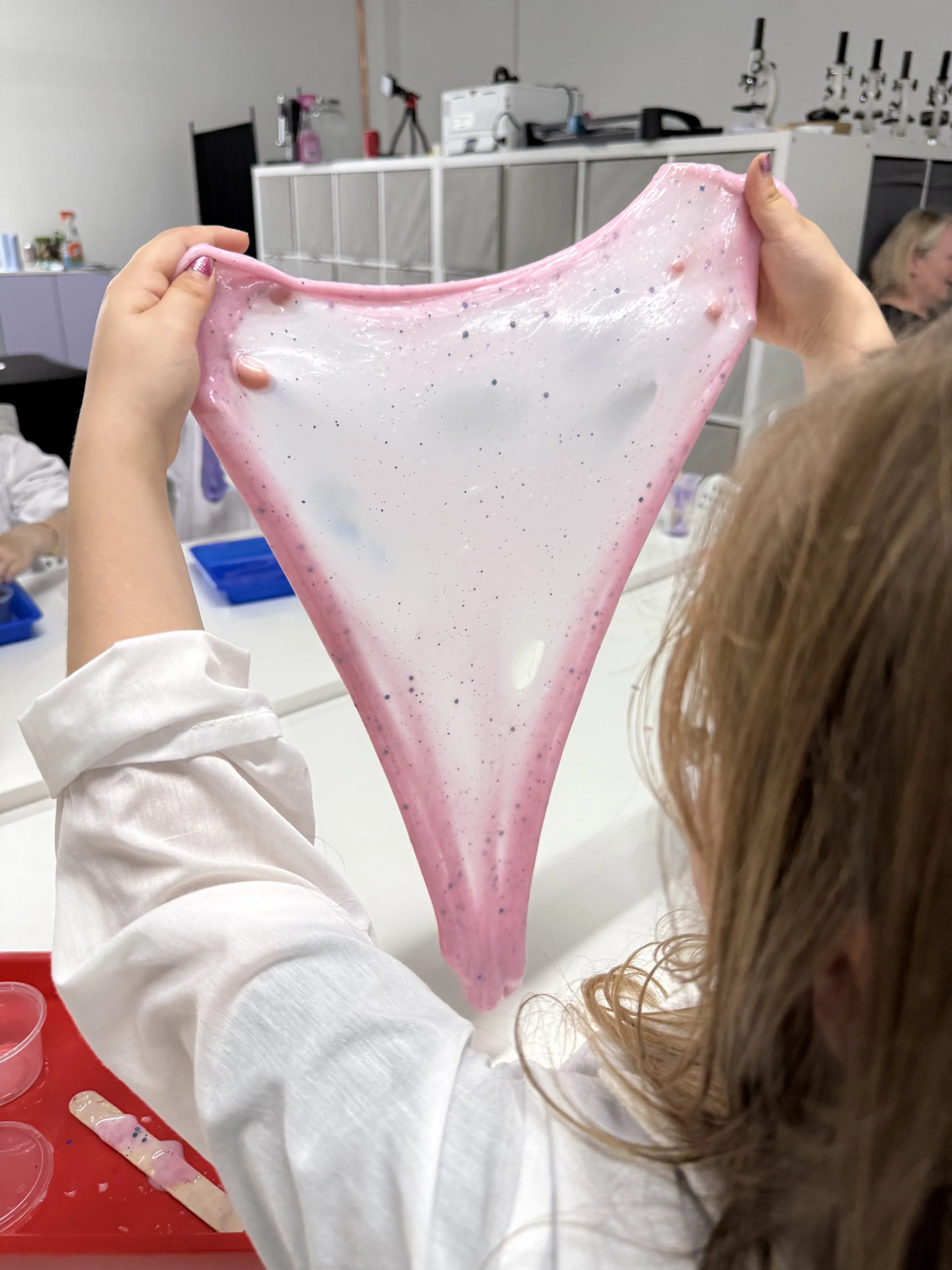 Person holding pink and white slime with blue and pink sparkles in a laboratory or classroom.