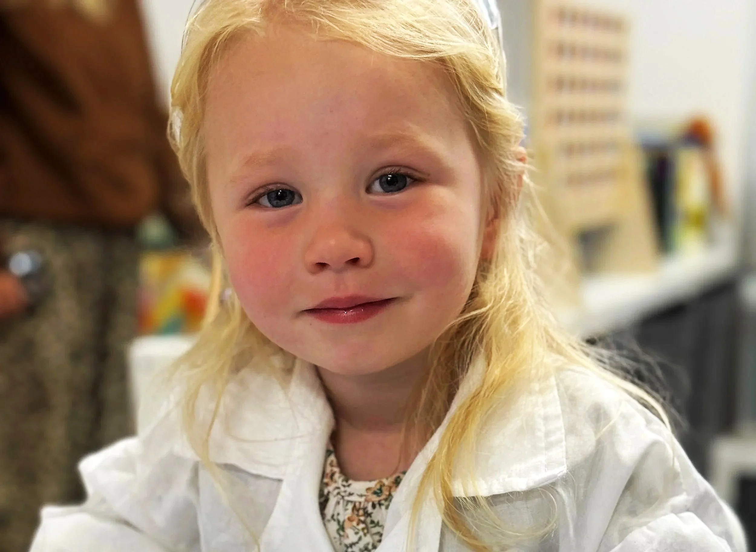 A young girl with blonde hair and blue eyes, smiling softly, wearing a white shirt with a floral pattern underneath, indoors with shelves and books in the background.