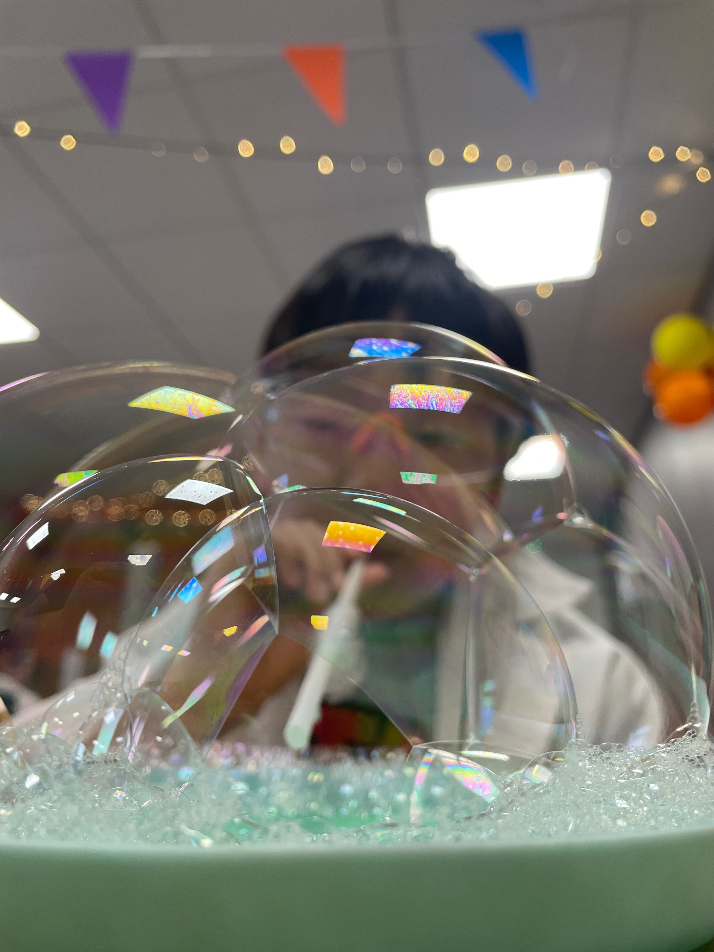Close-up view of iridescent soap bubbles floating above a container of bubbly soap foam, with a person making them in the background.