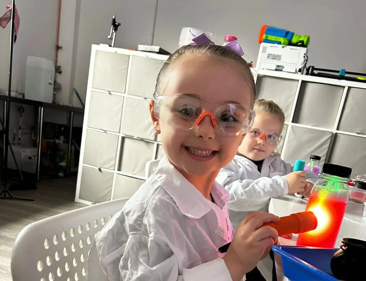 Two young girls wearing safety goggles and white lab coats engaging in a science experiment at a table, with a brightly lit container emitting red light.