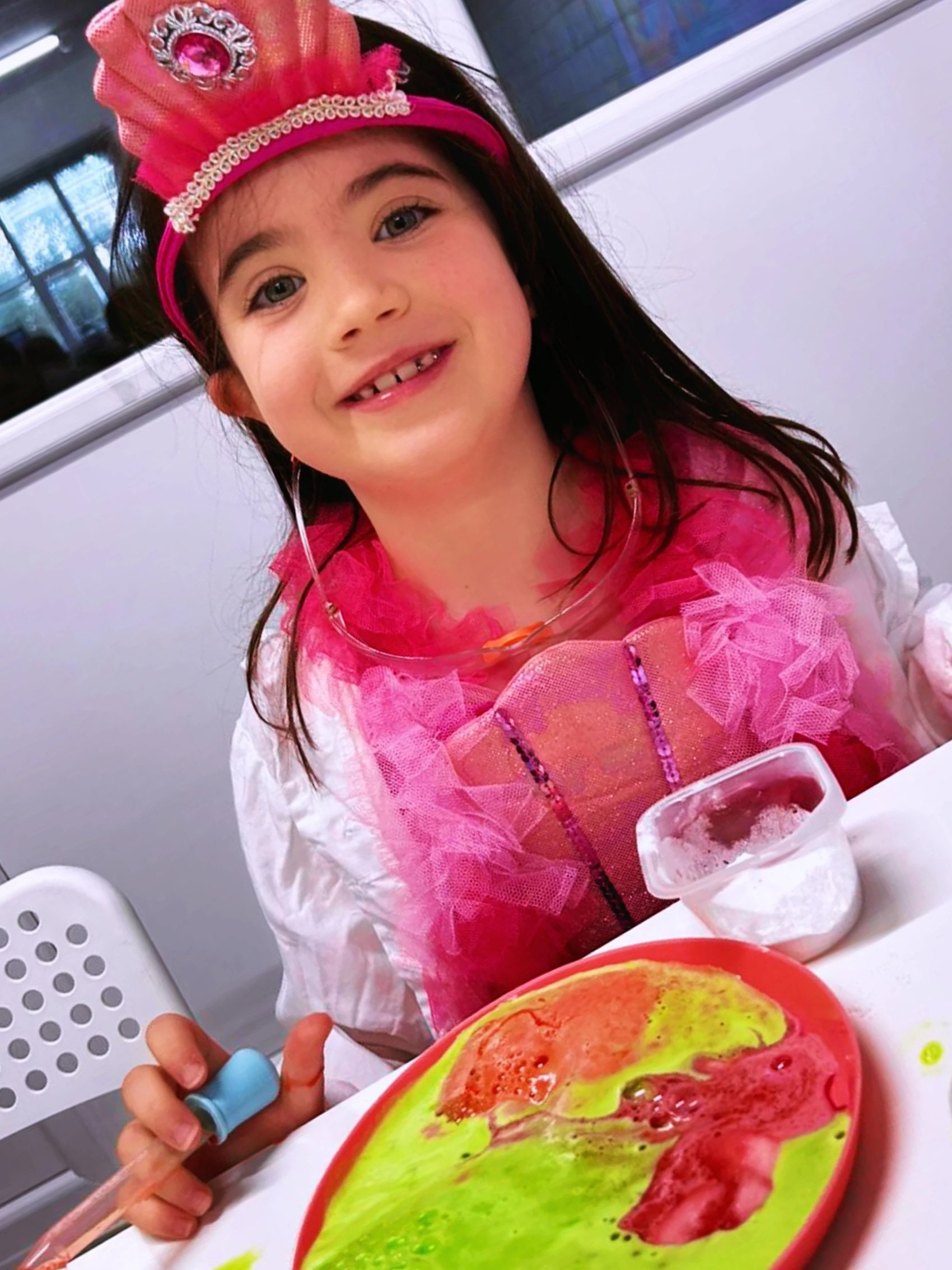 A young girl dressed in a princess costume with a pink crown and tutu, sitting at a table with a colorful plate of watermelon and greens, a small clear container with a beverage, and a plastic spoon, smiling at the camera.