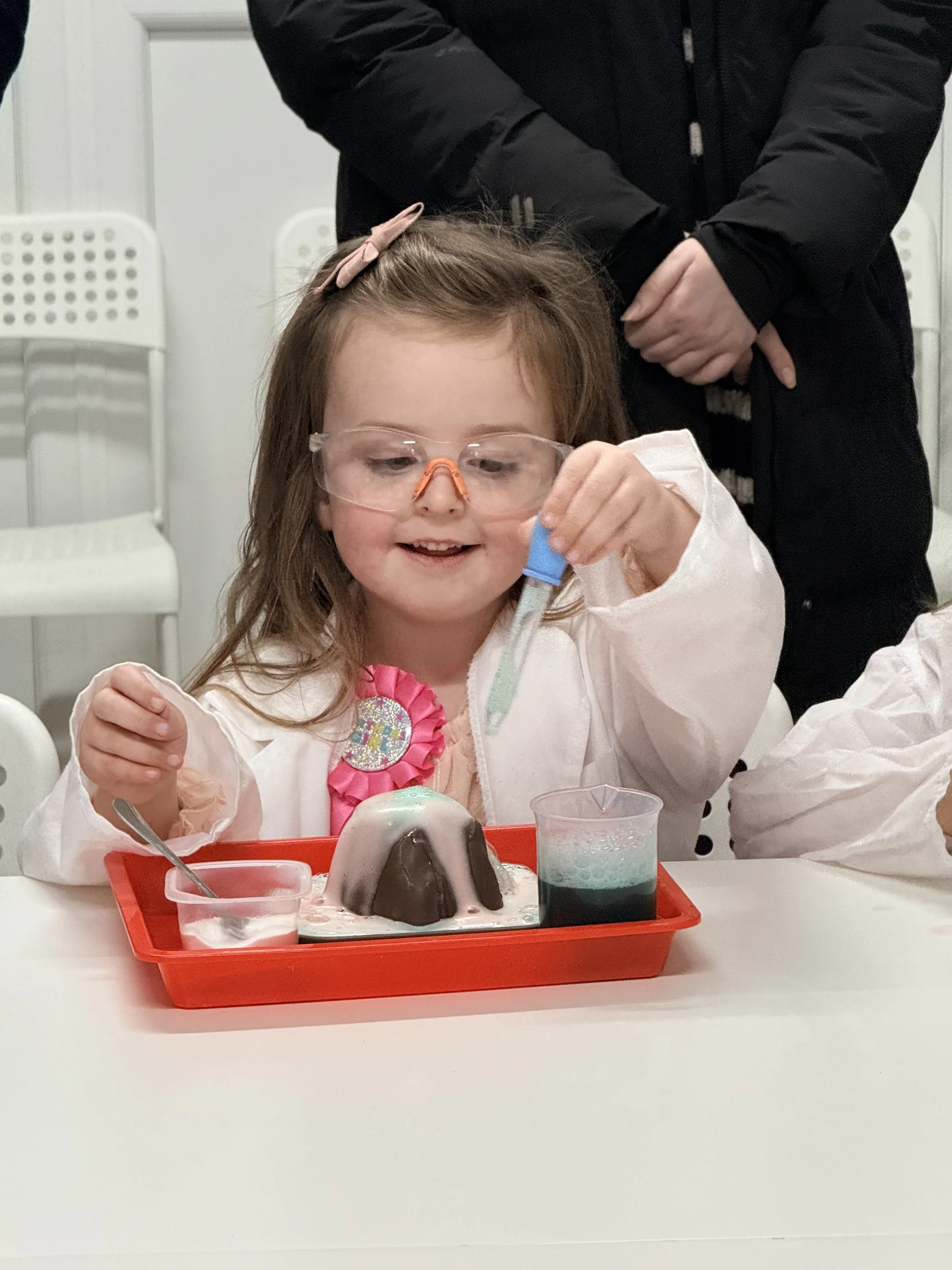Young girl wearing safety goggles and a white lab coat experimenting with science materials at a table, with a girl standing behind her. The girl is holding a test tube over a tray with a volcano cake and a beaker of colored liquid.