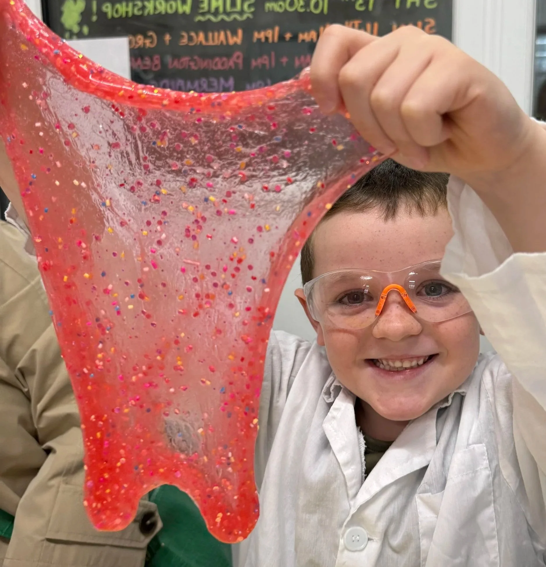 A young boy wearing safety glasses smiling while holding up a large piece of pink slime with colorful glitter.
