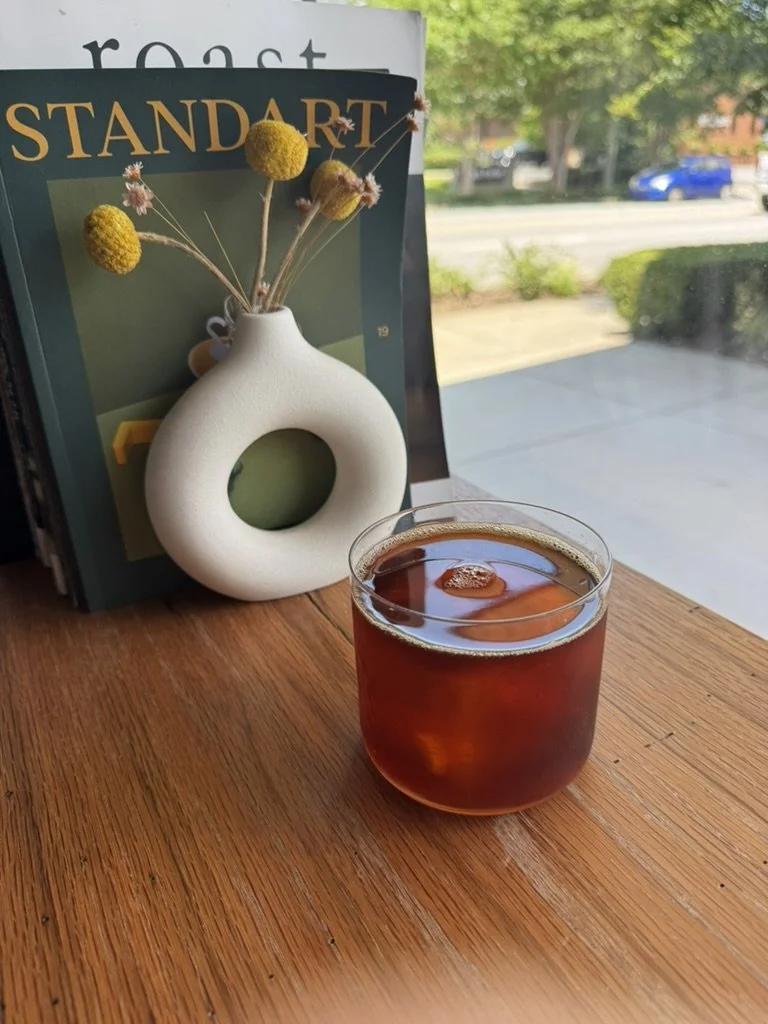 A glass of iced coffee on a wooden table, positioned near a white vase with yellow and pink flowers, and some books in the background near a window with a view of a street and trees.