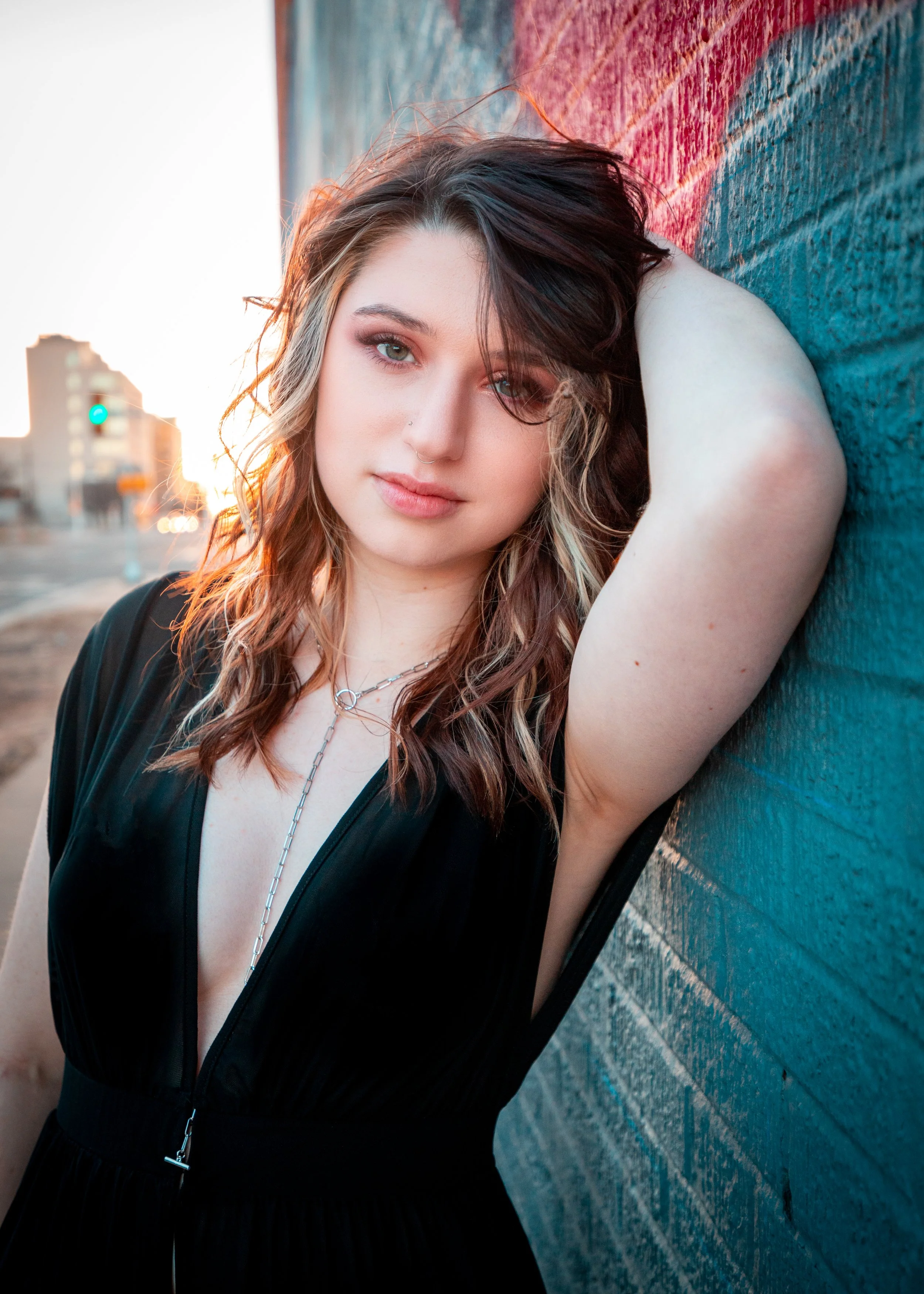 Young woman with wavy hair and makeup leaning against a brick wall at sunset, wearing a black zip-up dress and silver jewelry.
