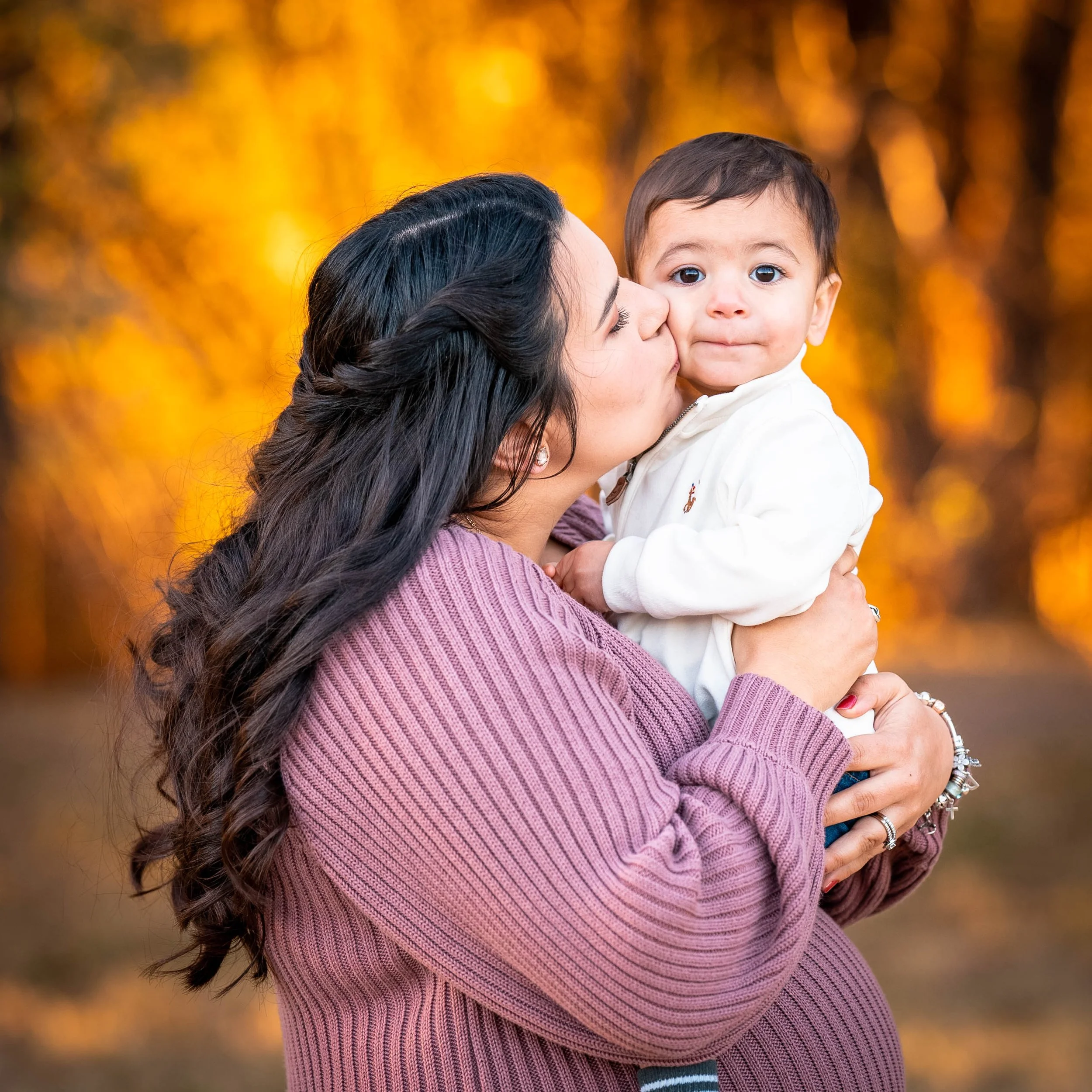 A woman with long dark hair wearing a mauve sweater is kissing a young boy on the cheek while holding him in her arms. The boy has short dark hair, big eyes, and is wearing a white jacket. The background features blurry autumn-colored trees.